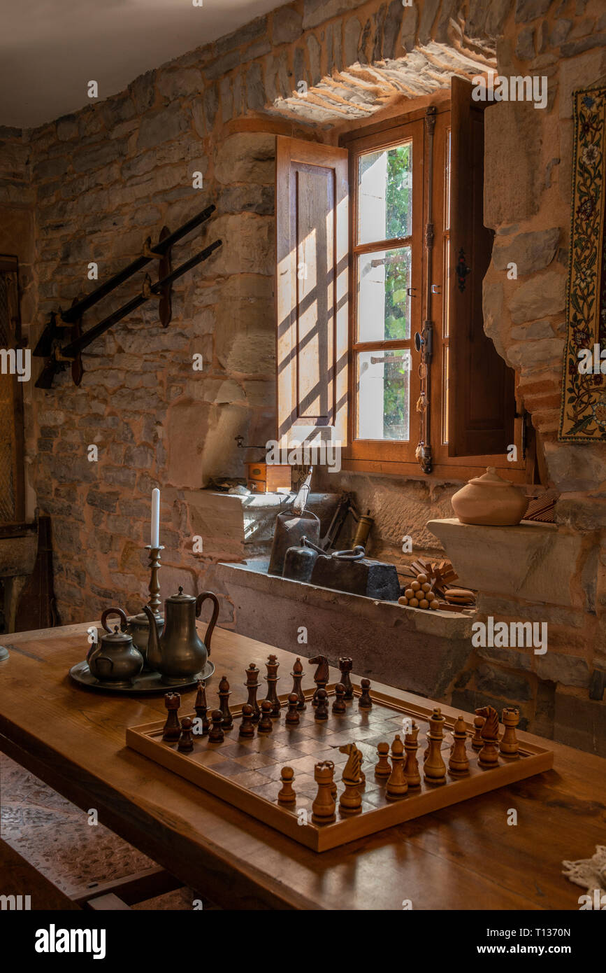 French country home interior, chess set and candle on a table, an old muskets mounted on the stone walls. All illuminated by sunlight through window. Stock Photo