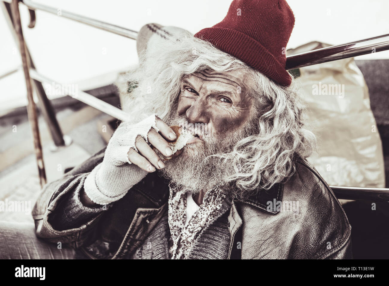 Homeless elderly eating roll that he buying for alms and looking on the camera. Stock Photo