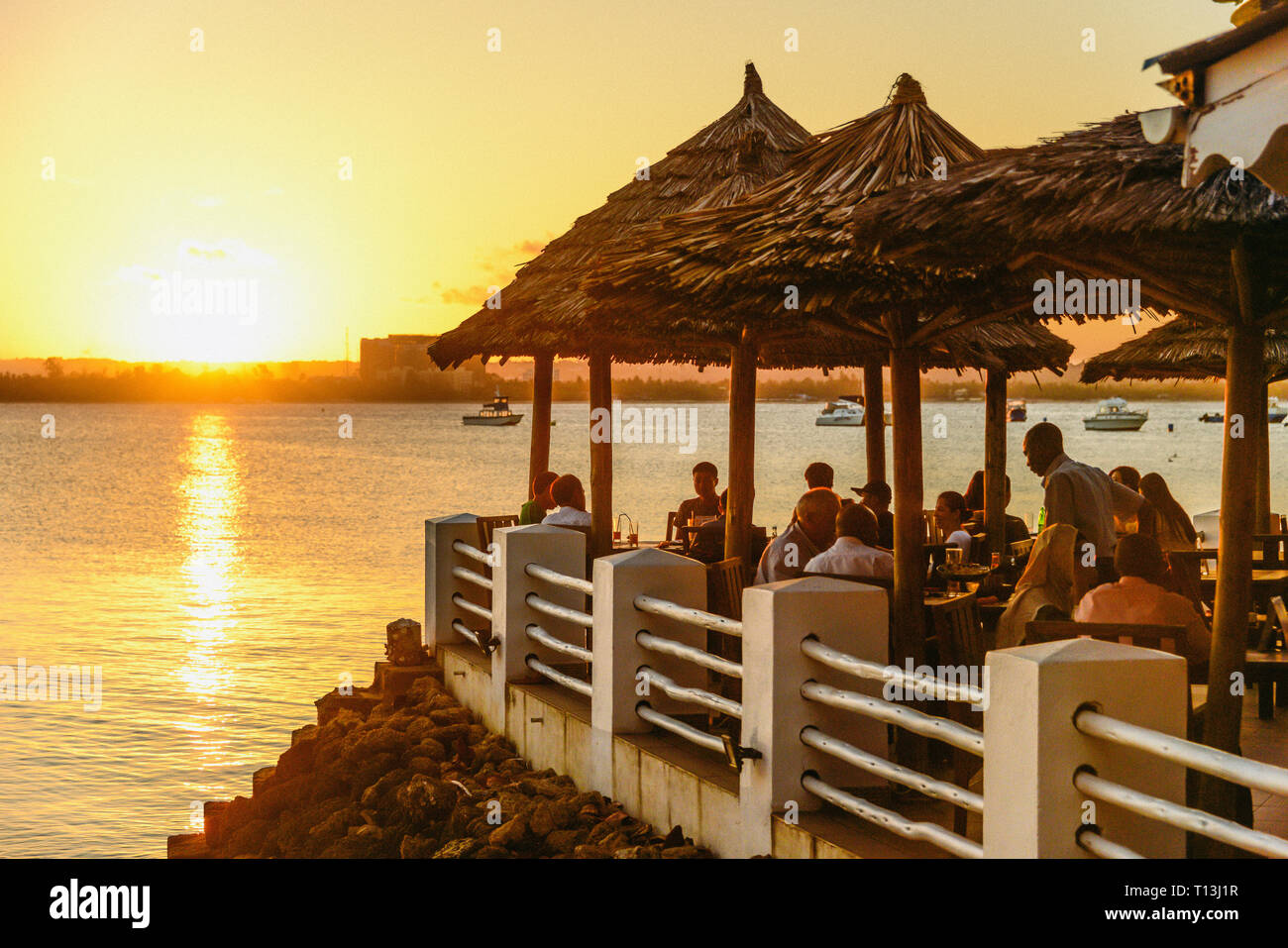 The setting sun backlights a beachside restaurant in Dar-es-Salaam, Tanzania. Palapa, or native reed umbrellas shelter the guests. Stock Photo