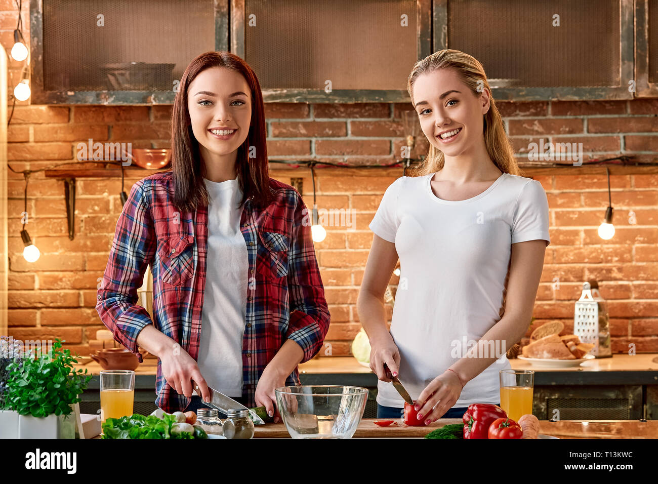 Portrait of two attractive women, having fun, while preparing vegetable salad. They are fully involved in the process. They are looking at the camera and smiling while slicing mushrooms. Front view Stock Photo