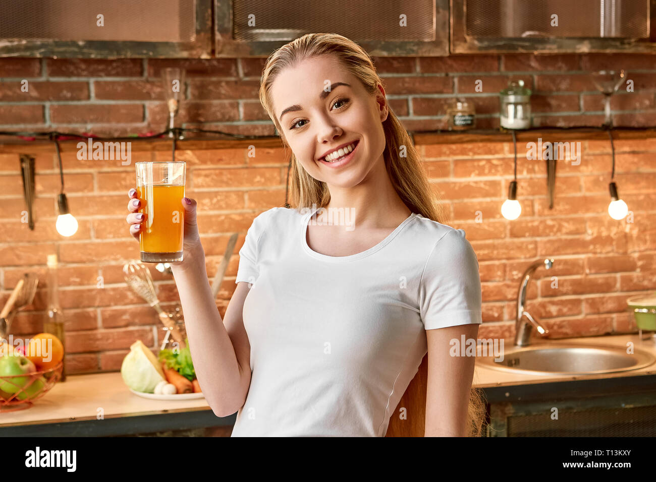 Portrait of cheerful blonde girl wearing white T-shirt, holding fresh juice, looking at the camera and smiling. Attractive young woman is preparing healthy food. Green healthy food concept. Front view Stock Photo