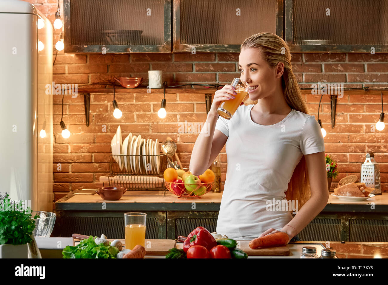 Portrait of blonde girl wearing white T-shirt, holding fresh juice, looking aside and smiling. Attractive young woman is preparing healthy food. Green healthy food concept. Vegetables and a glass of juice are on the tabletop. Front view. Stock Photo