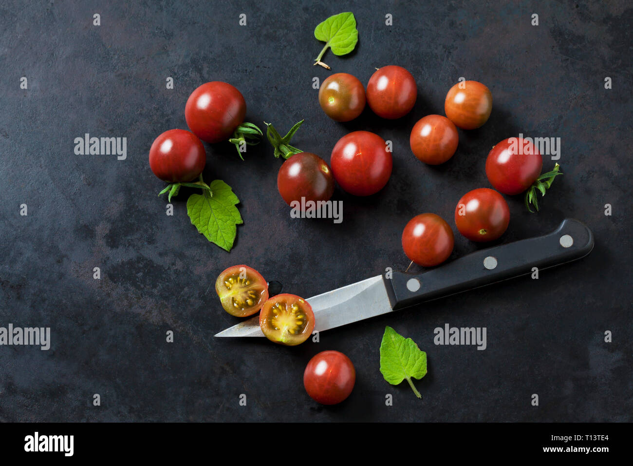 Whole and sliced risp tomatoes 'Black Cherry', leaves and kitchen knife on dark ground Stock Photo