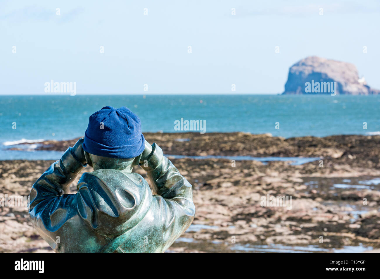 North Berwick, East Lothian, Scotland, UK, 23rd March 2019. UK Weather: Sunny Spring weather at the seaside town. Bronze statue called The Watcher by Kenny Hunter with a warm hat as he surveys Bass Rock gannet colony through binoculars Stock Photo