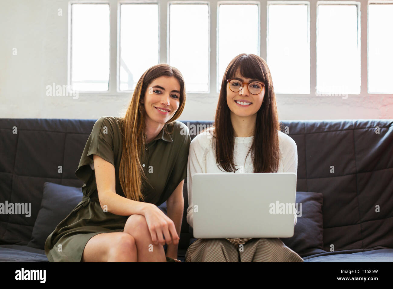 Two smiling young women sitting on couch in office with laptop Stock Photo