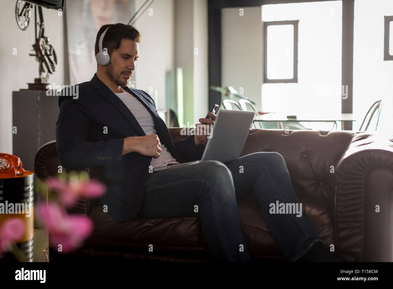 Man with headphones sitting on couch using cell phone and laptop Stock Photo