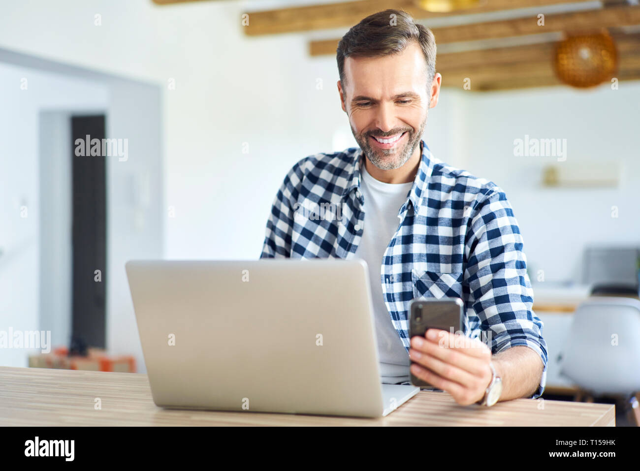 Cheerful man using phone while working on laptop at home Stock Photo