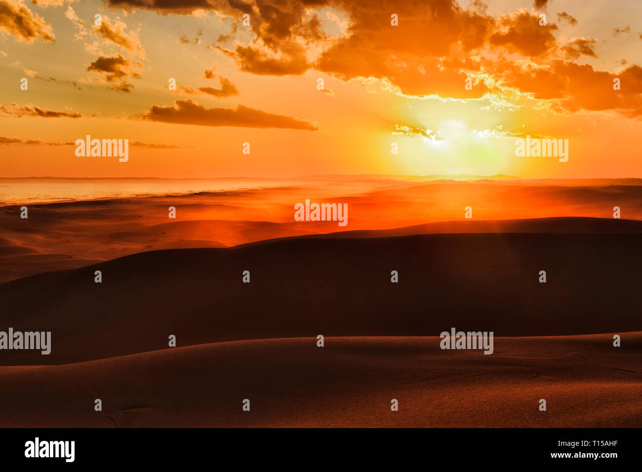 The sun setting over horizon behind long deserted sand dunes of Stockton beach on Pacific coast of Australia - bright orange sunlight hues sands and s Stock Photo