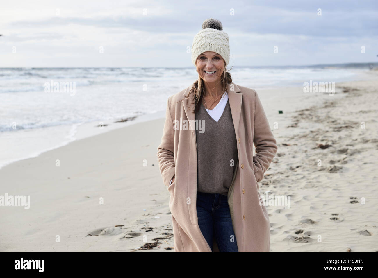 Spain, Menorca, portrait of smiling senior woman wearing bobble hat and coat on the beach in winter Stock Photo