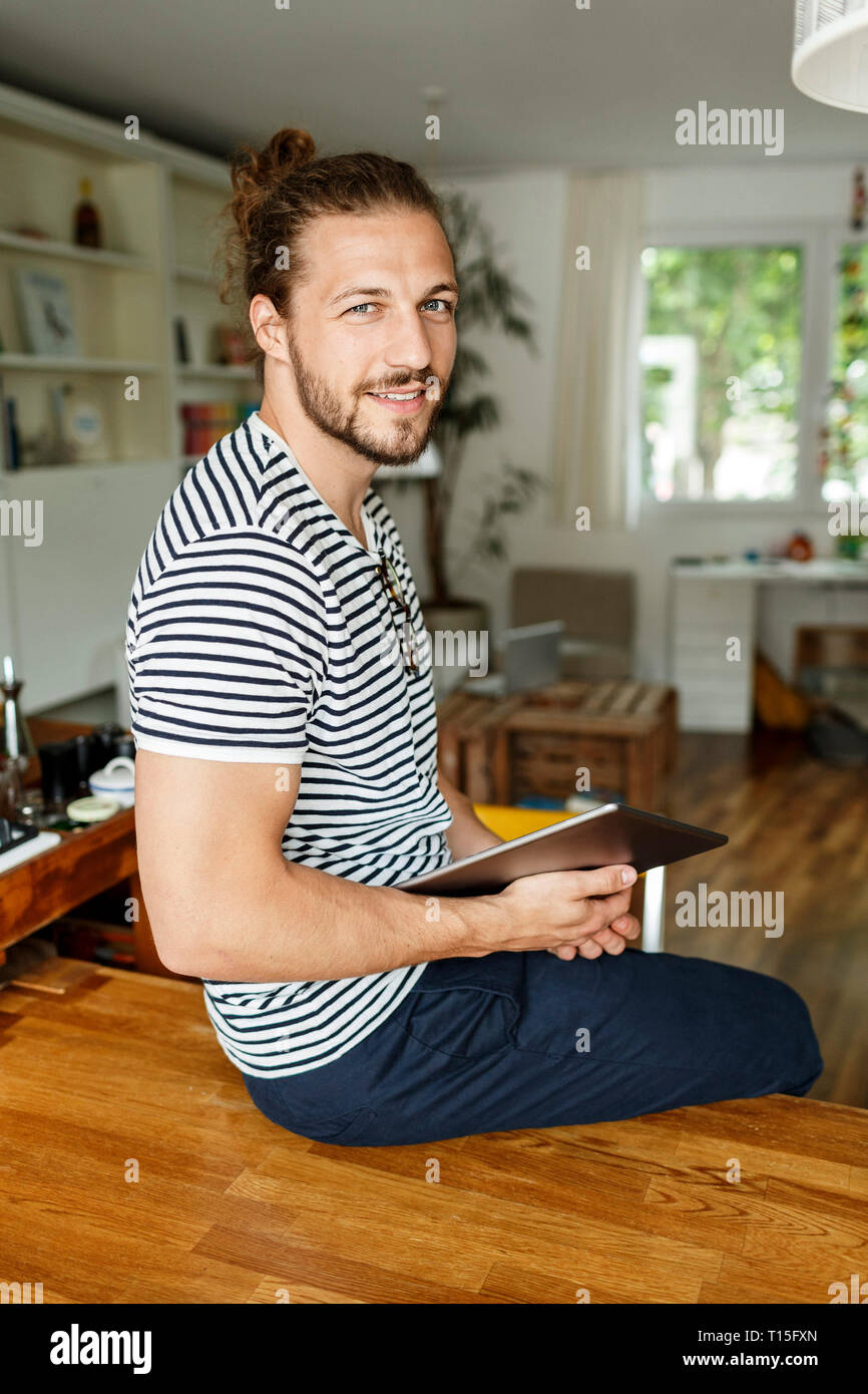 Young man with a bun sitting on table, using digital tablet Stock Photo