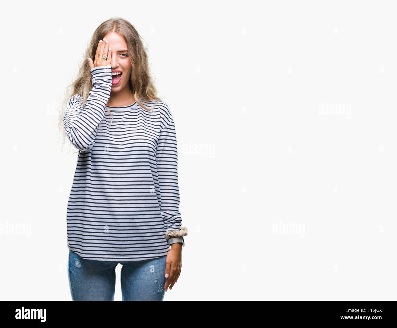 Beautiful young blonde woman wearing stripes sweater over isolated background covering one eye with hand with confident smile on face and surprise emo Stock Photo
