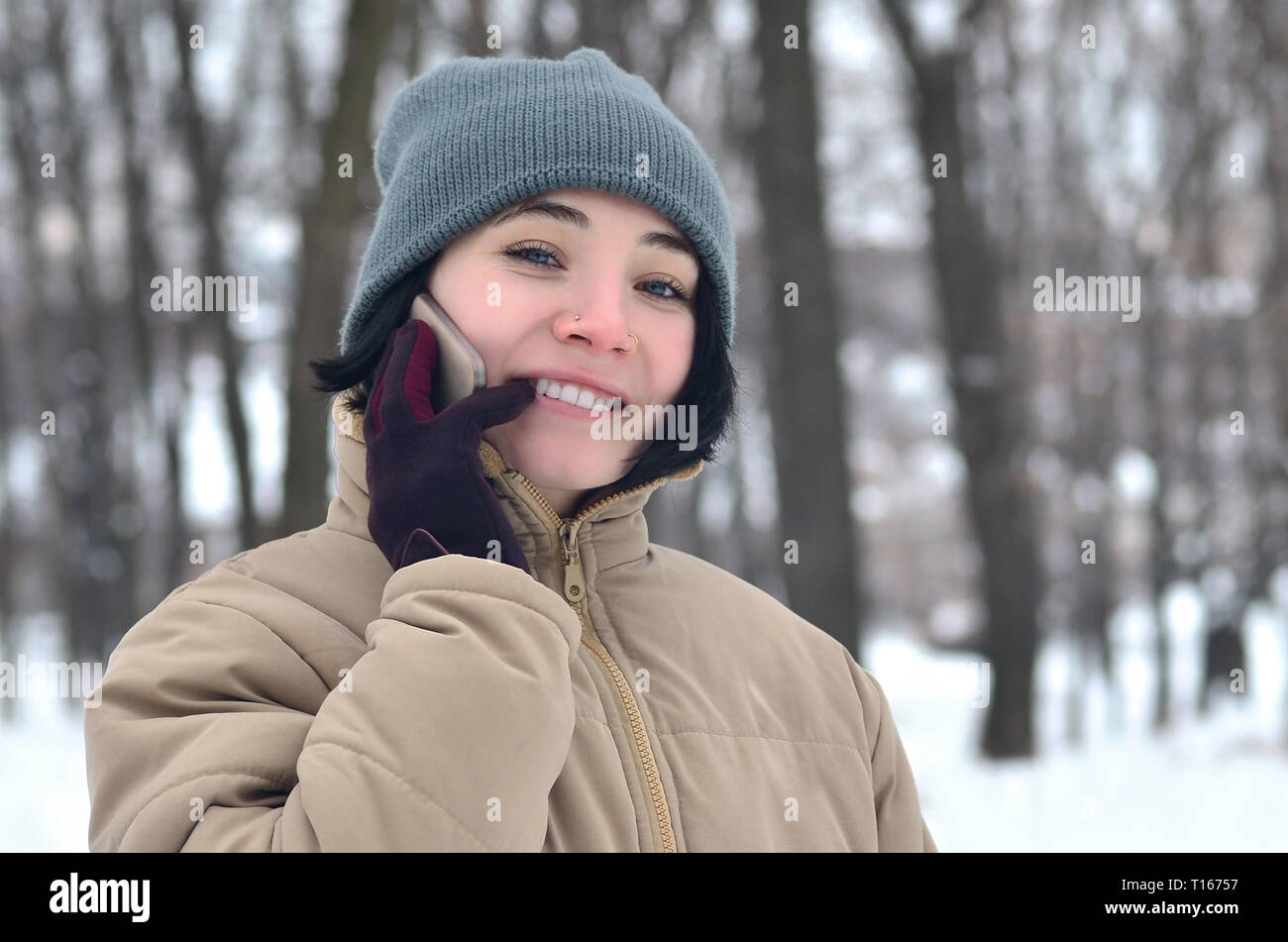 Portrait of pretty attractive caucasian pierced girl hipster in winter park. She is talking on modern smartphone. Youth street fashion. Winter fun. Wa Stock Photo