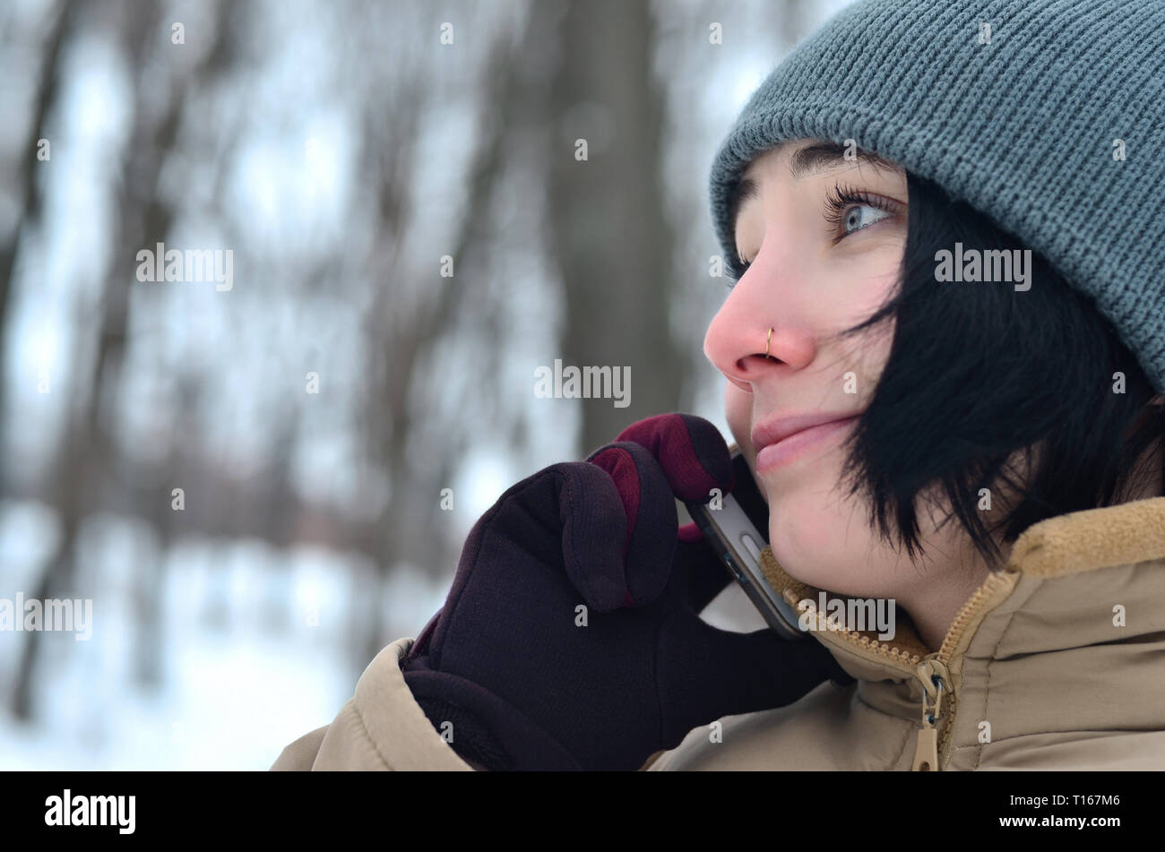 Beauty girl in winter forest. Beautiful young brunette caucasian pierced woman using smartphone in fashionable brown coat and warm grey hat Stock Photo