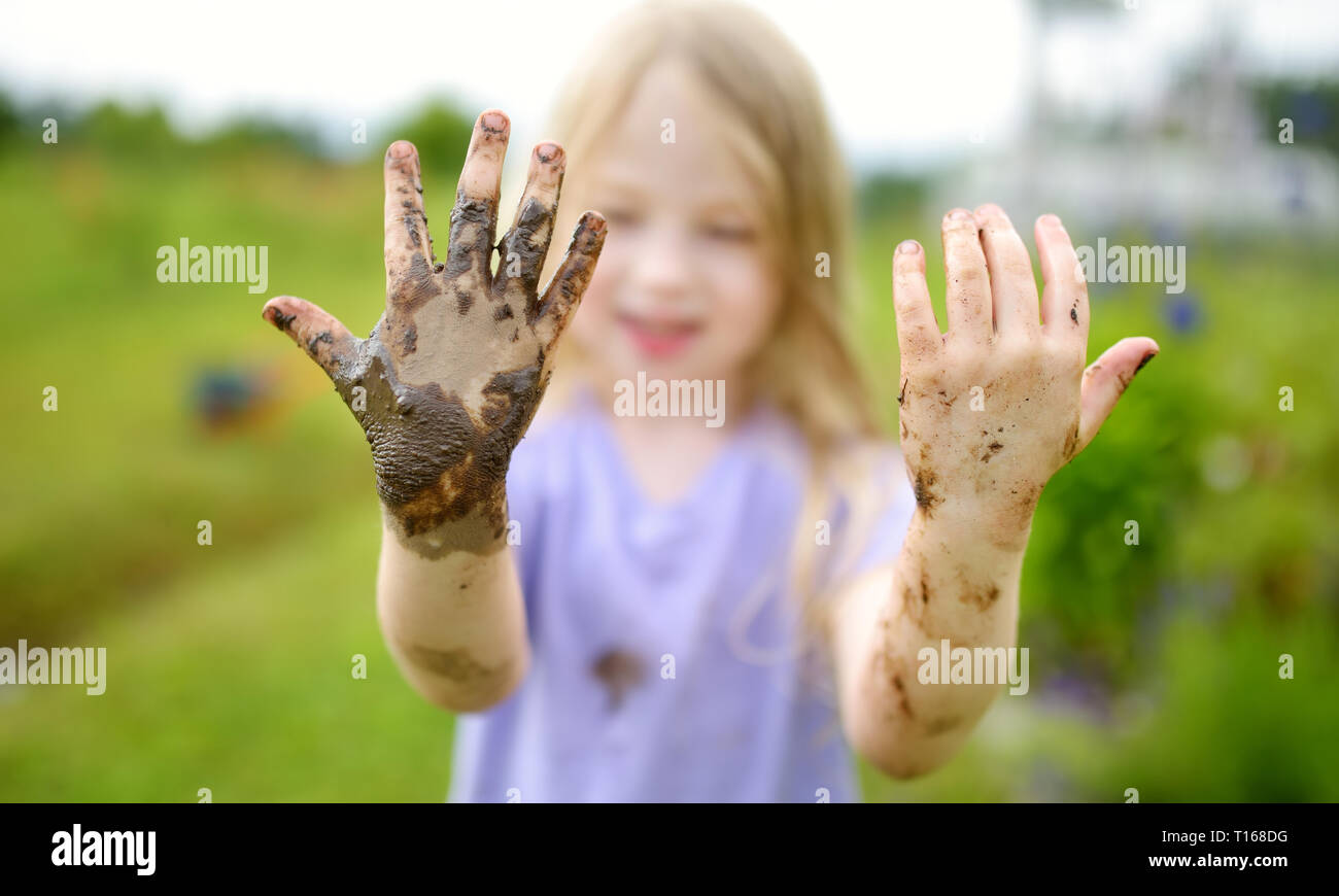 Funny little girl playing in a large wet mud puddle on sunny summer day. Child getting dirty while digging in muddy soil. Messy games outdoors. Stock Photo