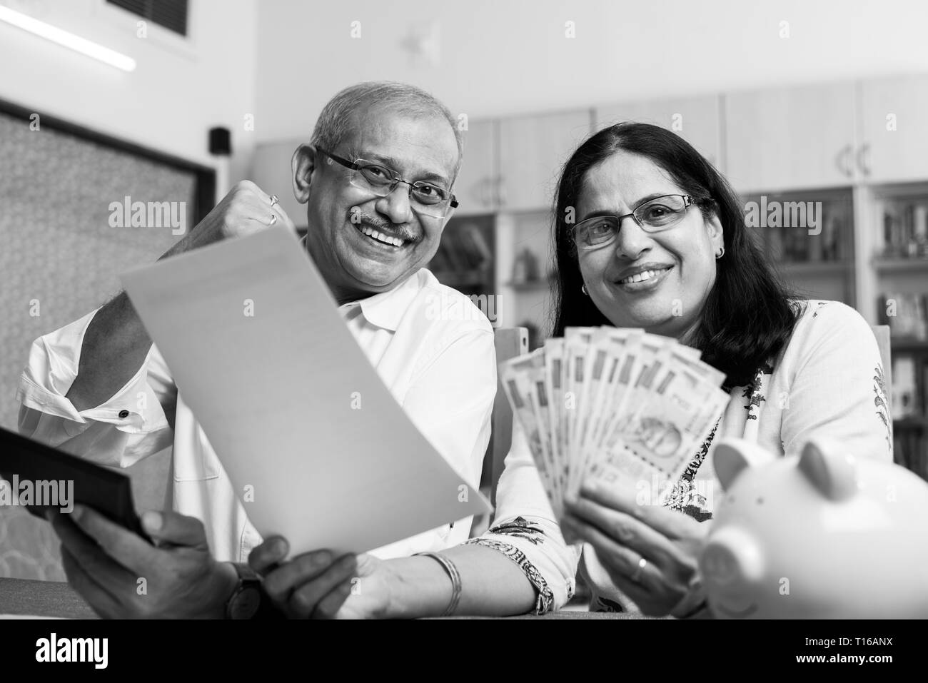 Senior Indian/asian couple accounting, doing home finance and checking bills with laptop, calculator and money while sitting on sofa/couch at home Stock Photo