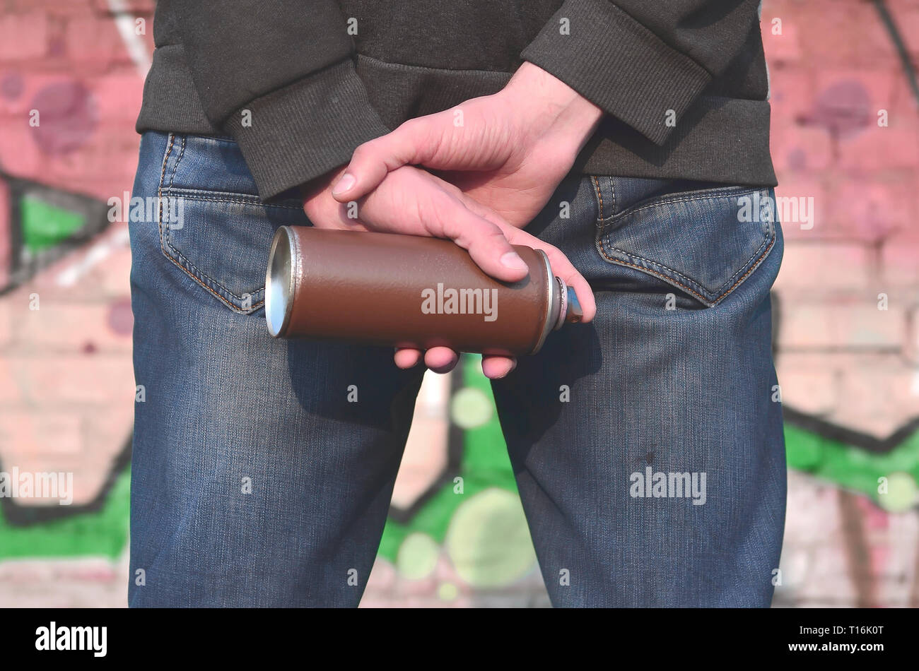 Rear view of the body of a young guy in blue dirty jeans and a gray sweater with a used paint spray in his hand against the background of an old brick Stock Photo