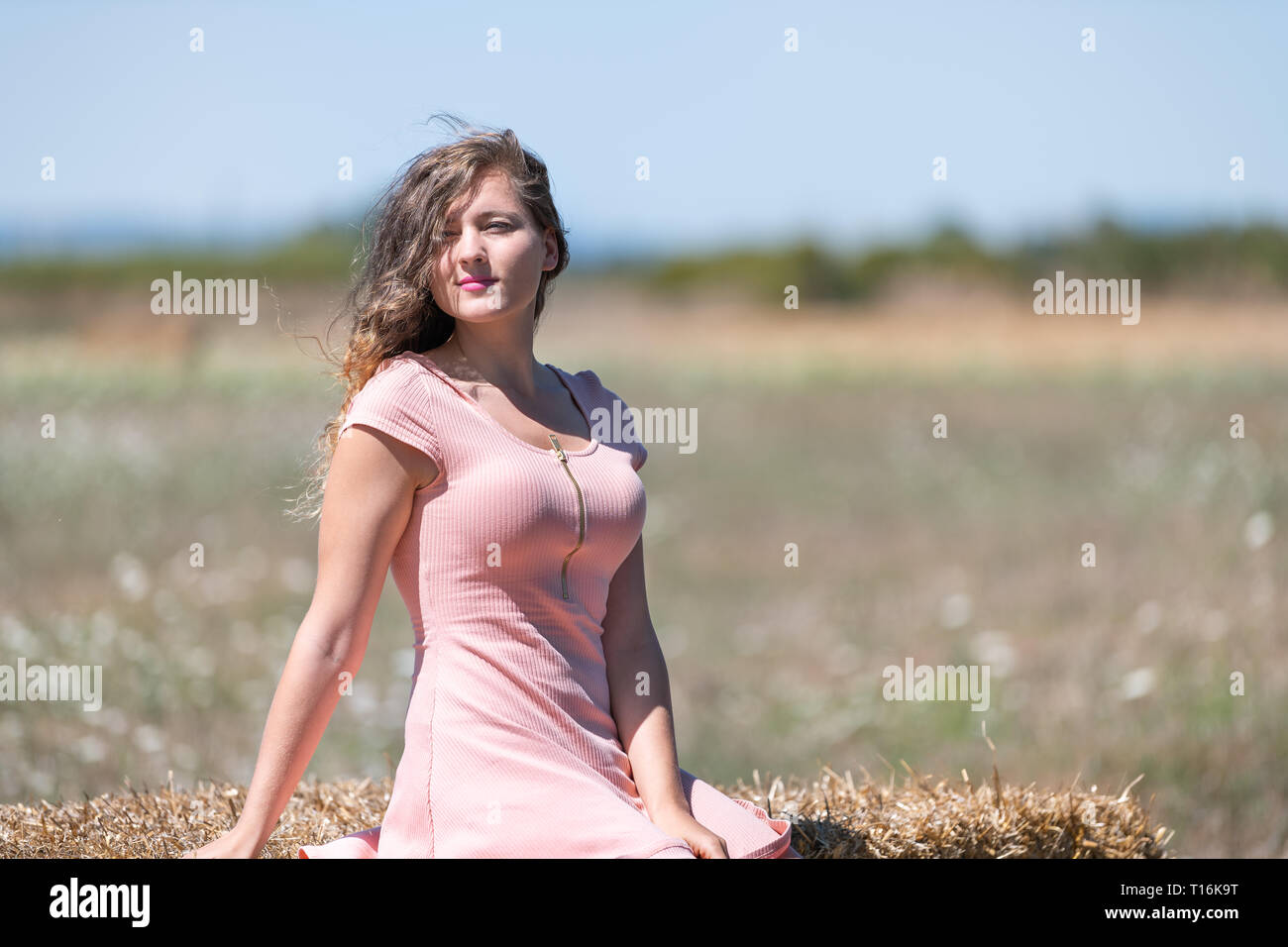 Countryside landscape in Tuscany, Italy with young girl woman sitting on hay bale in dress with hair in wind posing model Stock Photo