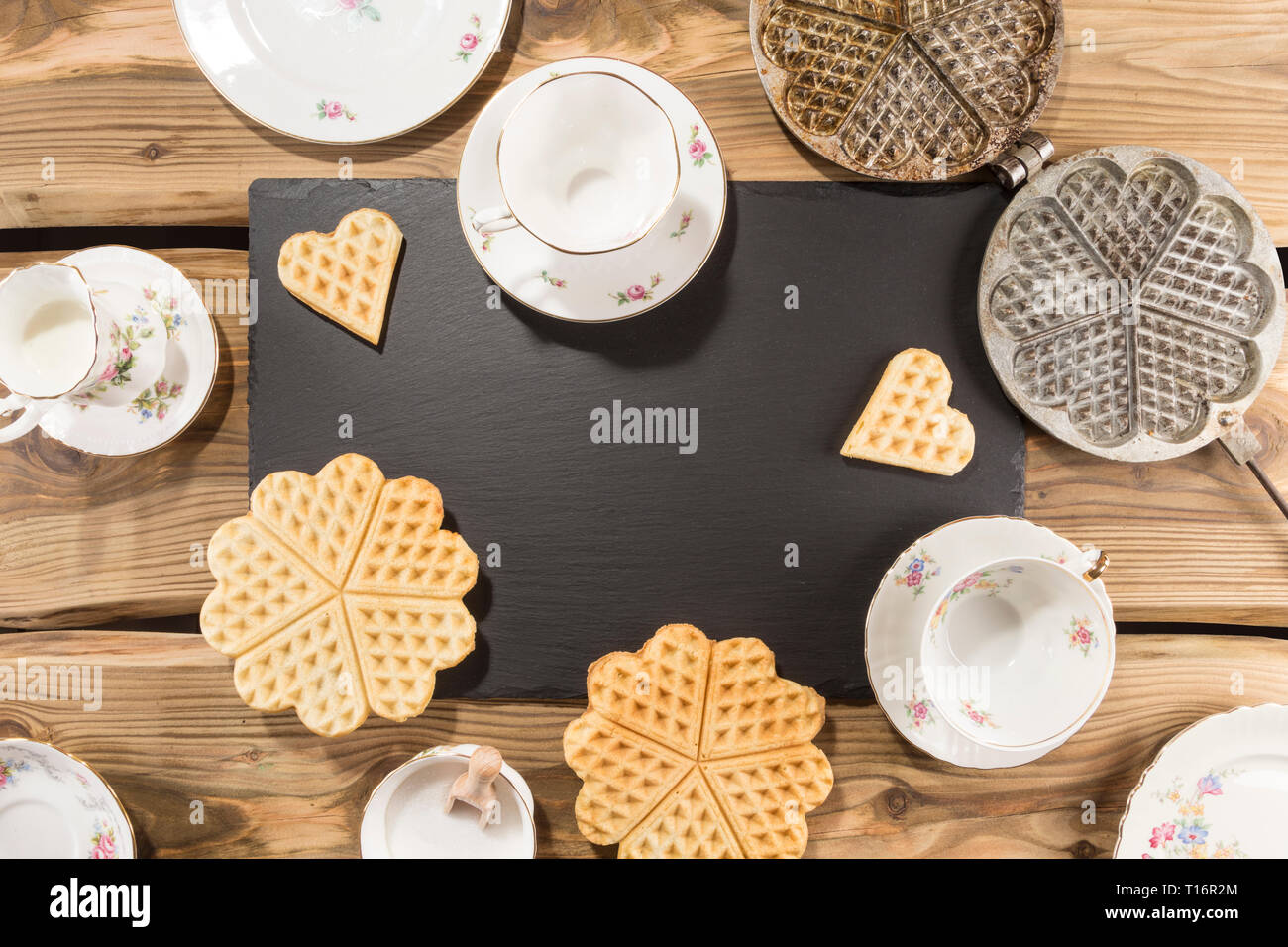 Delicious waffles, traditional ancient waffle iron arranged on rustic wood planks with slate board and English porcelain. Shot from above, flat lay. Stock Photo