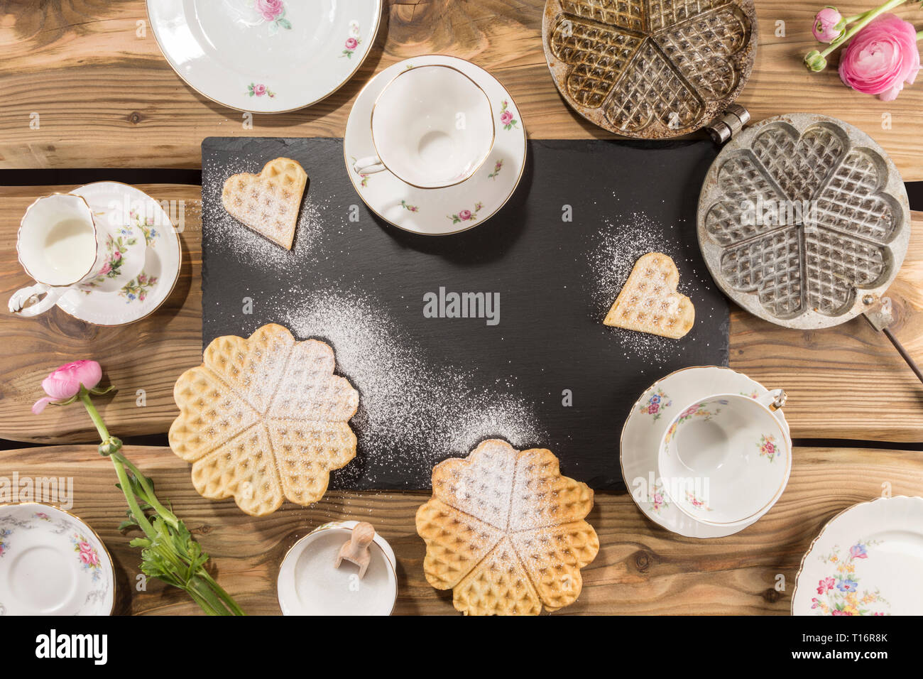 Delicious waffles, traditional ancient waffle iron arranged on rustic wood planks with slate board and English porcelain. Shot from above, flat lay. Stock Photo