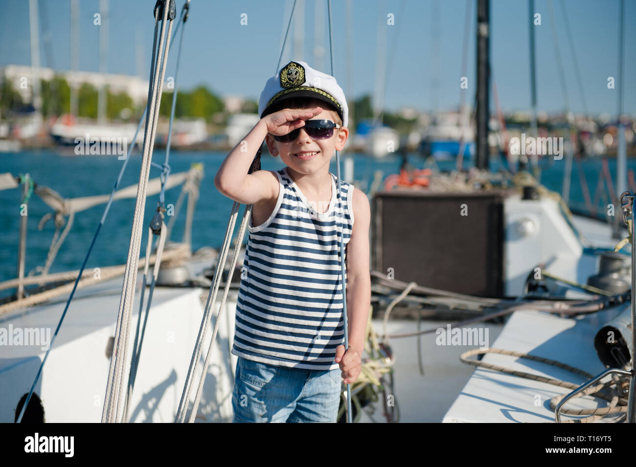 cute-happy-kid-in-captain-cap-on-yacht-board-in-summer-sea-port-T16YT5.jpg