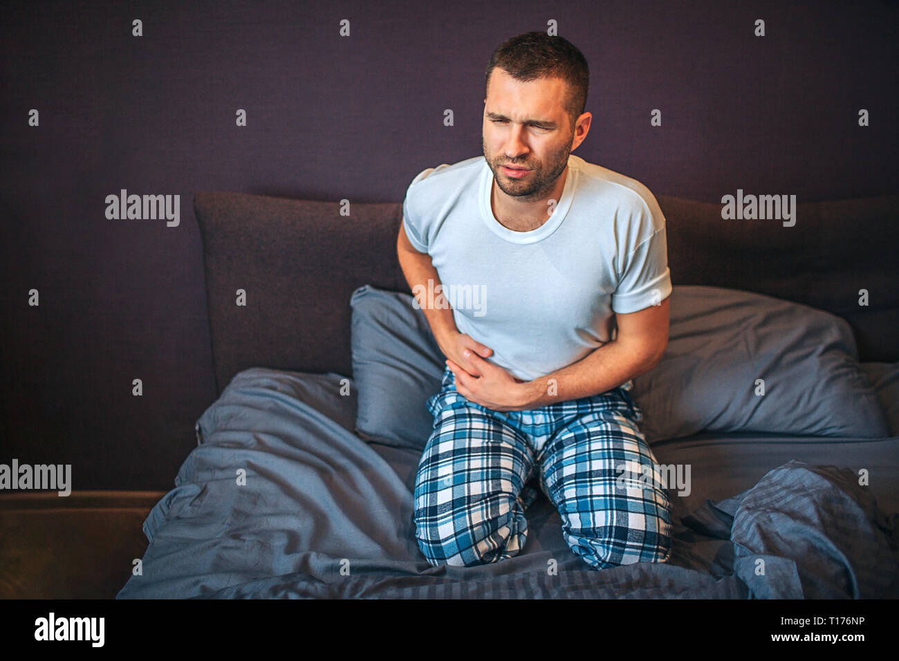 Young man stands on knees on bed and suffers from pain in appendix area. He holds hands there. Guy shrinks. He wear pajamas. Stock Photo
