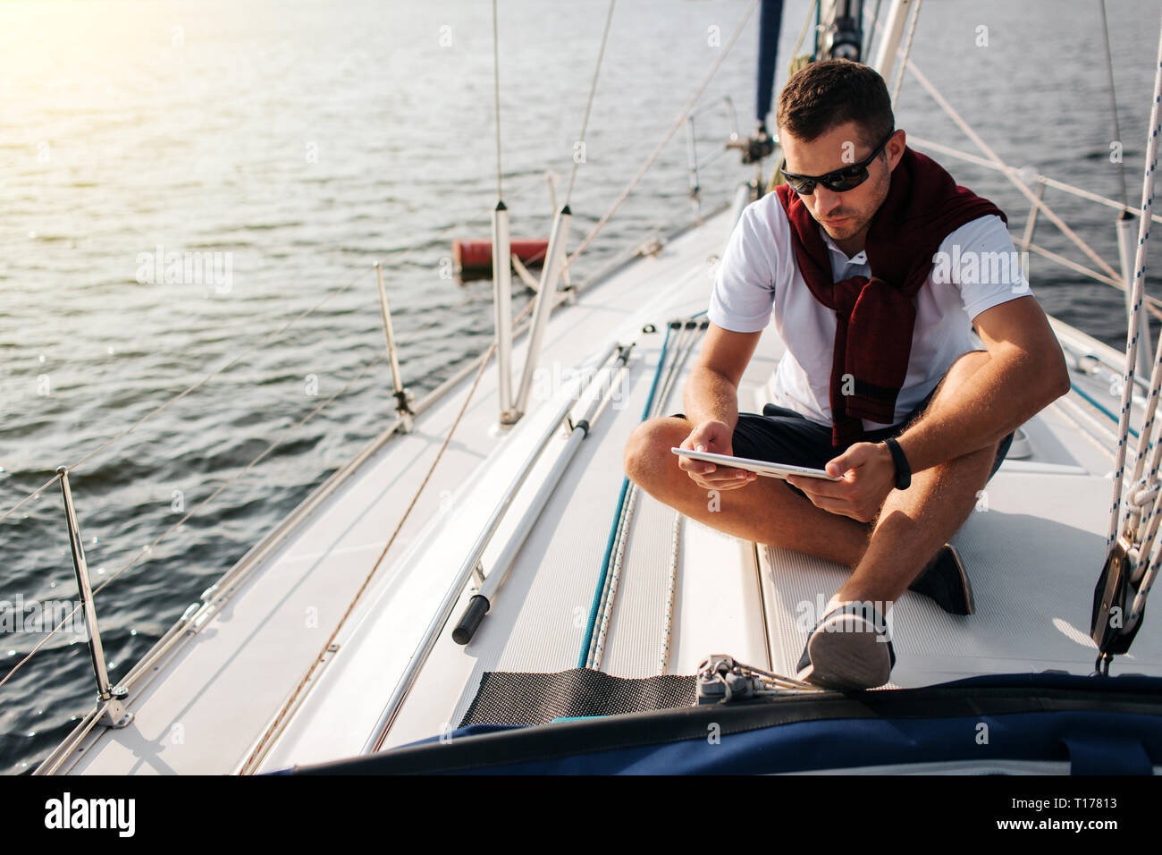 Serious and peaceful guy sits on board of yacht. He holds and looks at tablet. Young man is calm. He wears white sirt and dark sweater with shorts. Stock Photo
