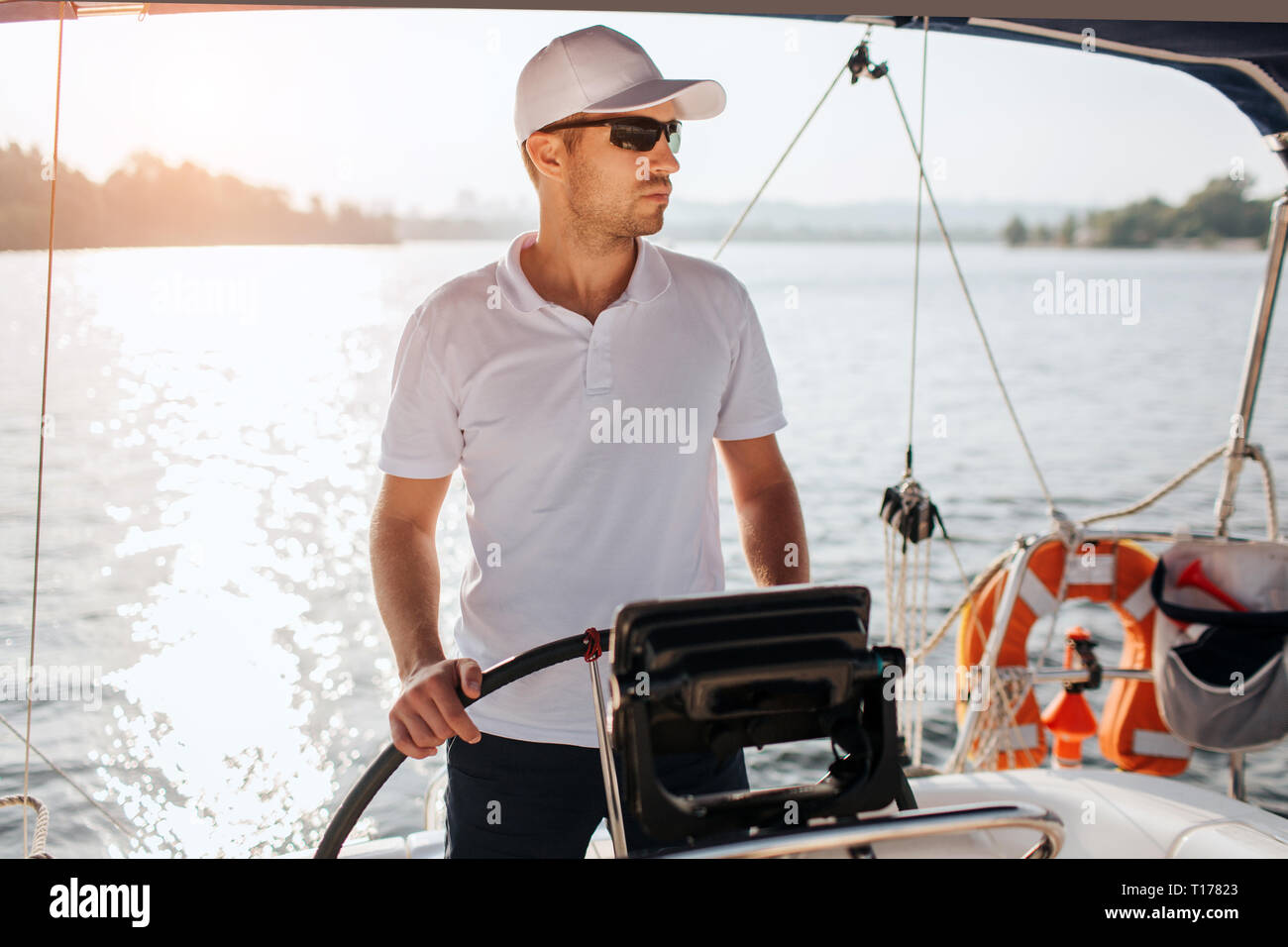 Prefessional sailor stands at yacht rudder and hold it with hands. He looks to right. Guy wear cap, sunglasses and white sirt with black shorts. It is Stock Photo