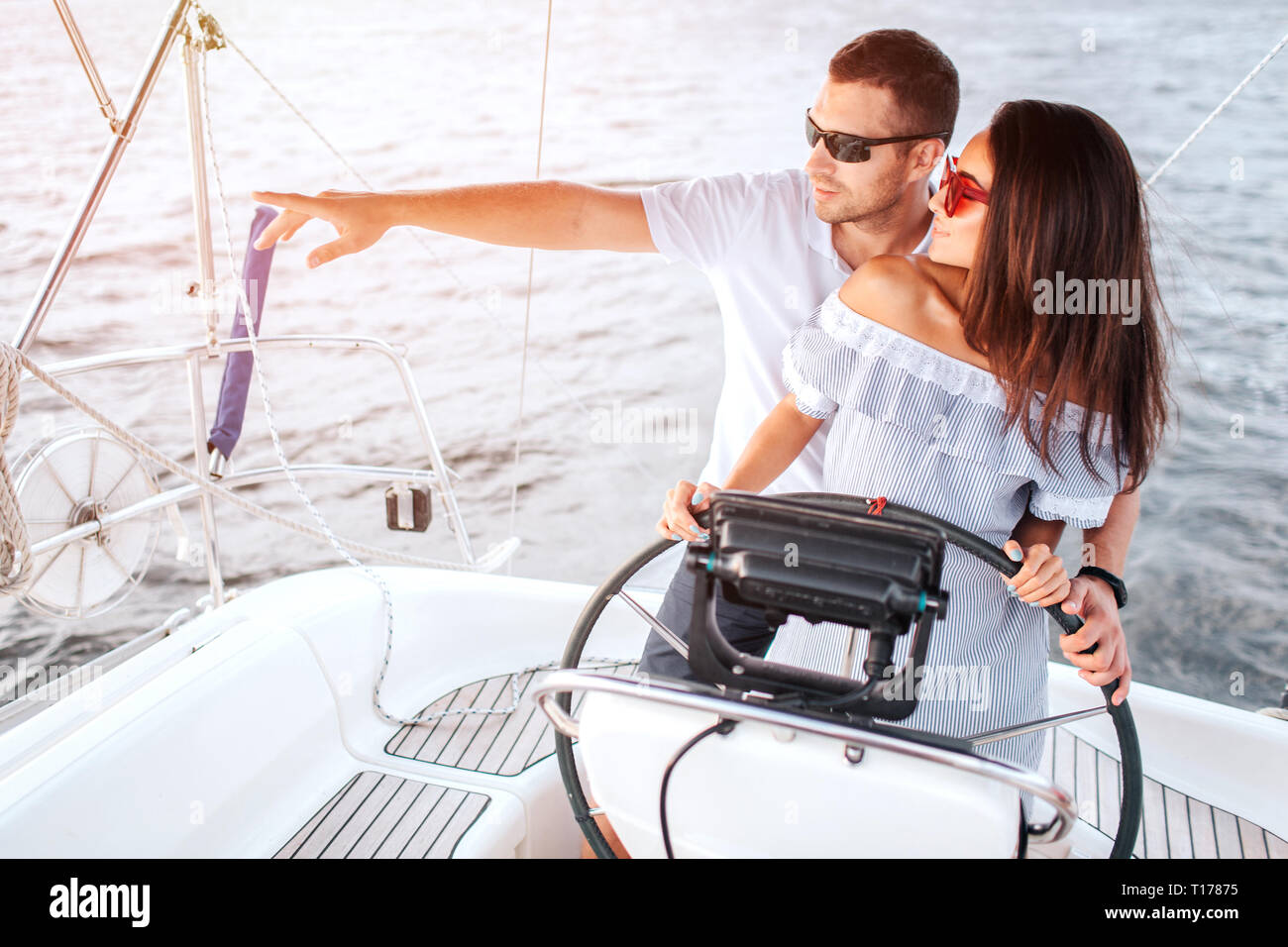 Nice picture of young people standing together and looking to left. He points with hand. She holds her hands on rudder. They wear sunglasses. Stock Photo