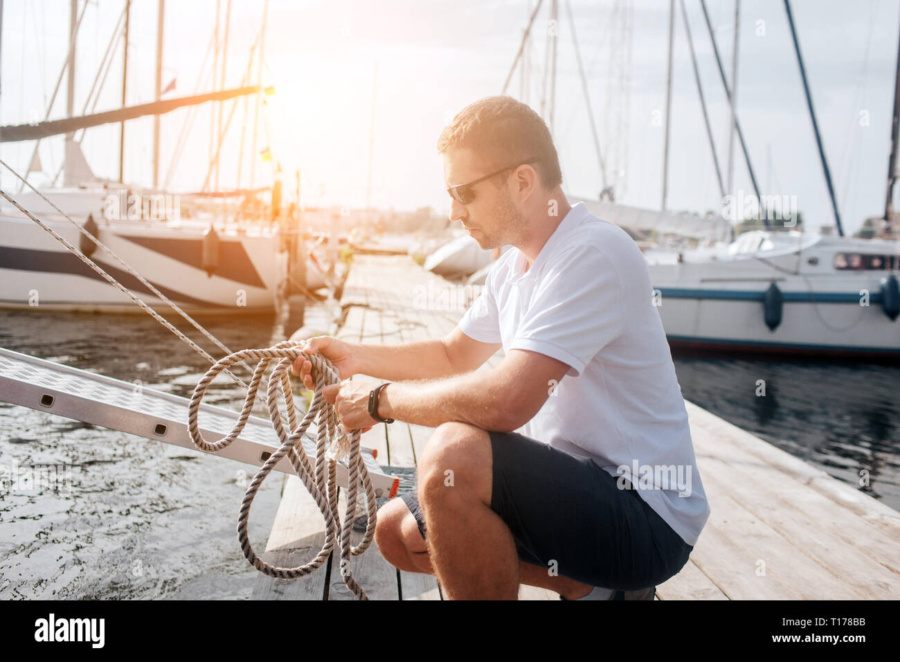 Strong and well-built sailor sits in squad position and holds ropes. He looks at them. Guy is tying knot. Stock Photo