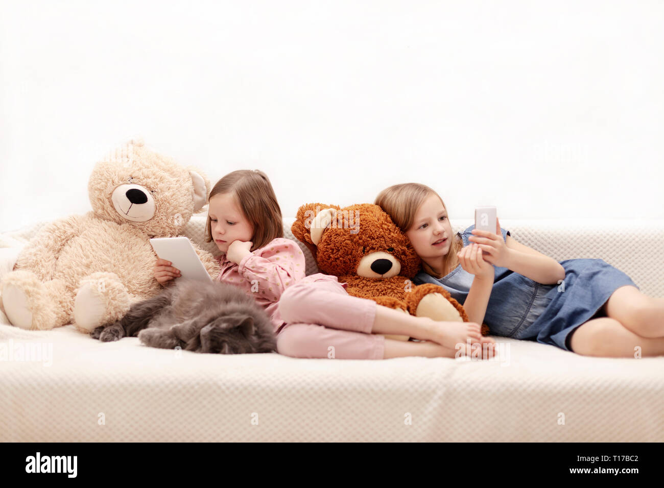 Two little girls (sisters 7 and 8 years old) are watching an e-book and a smartphone at home on the sofa with their cat. Selective focus. Stock Photo