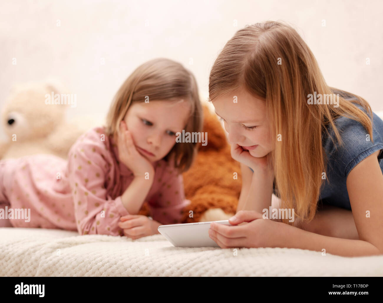 Two little girls (sisters 7 and 8 years old) are watching an e-book and a smartphone at home on the sofa with their cat. Selective focus. Stock Photo