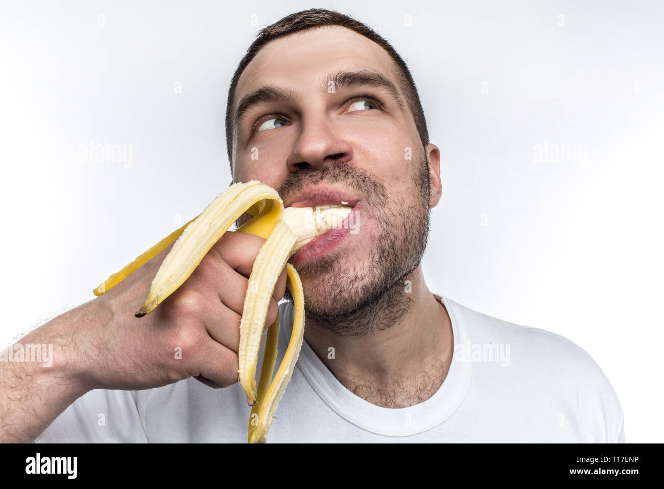 Another strange picture of unshaved guy eating ripe banana. He is biting a big piece of fruit. Man is enjoying the mmoment and looking somewhere aside Stock Photo