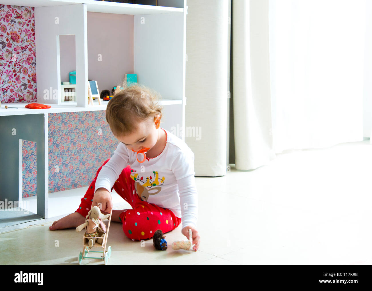 backlit image of toddler playing with doll house and stuffed animal toys, sitting on tiled,flooring at home alone with pacifier in mouth. Stock Photo