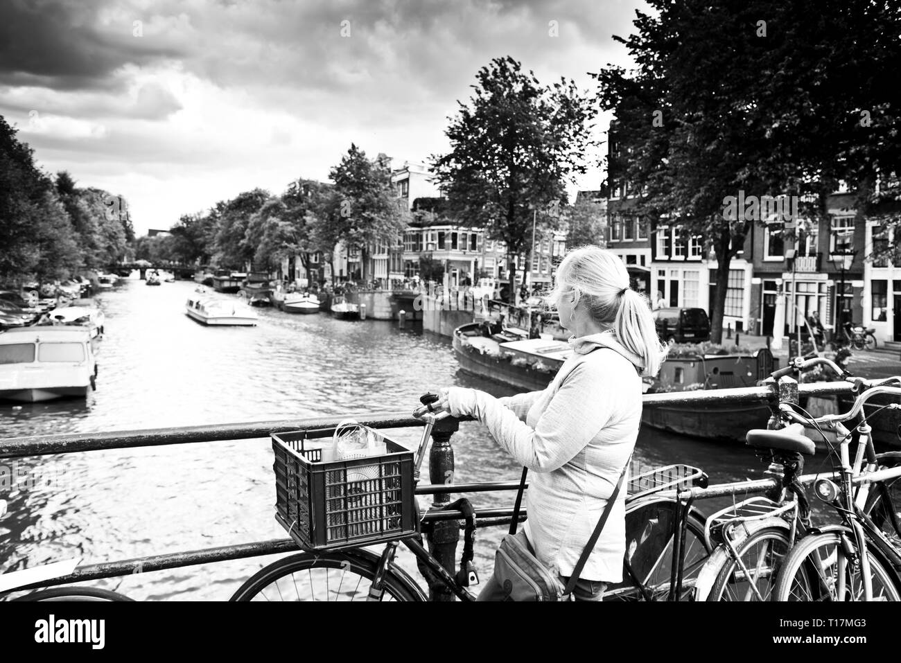 woman stand next to her parked bicycle with shopping in her basket in the middle of a bridge watching canal traffic passing,stormy sky above. Stock Photo