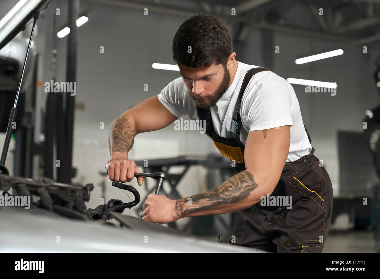 Car repair and maintenance. Young muscular mechanic in coveralls and white t shirt repairing car in autoservise station. Handsome bearded worker with dark hair and tattoos on hands holding tool. Stock Photo