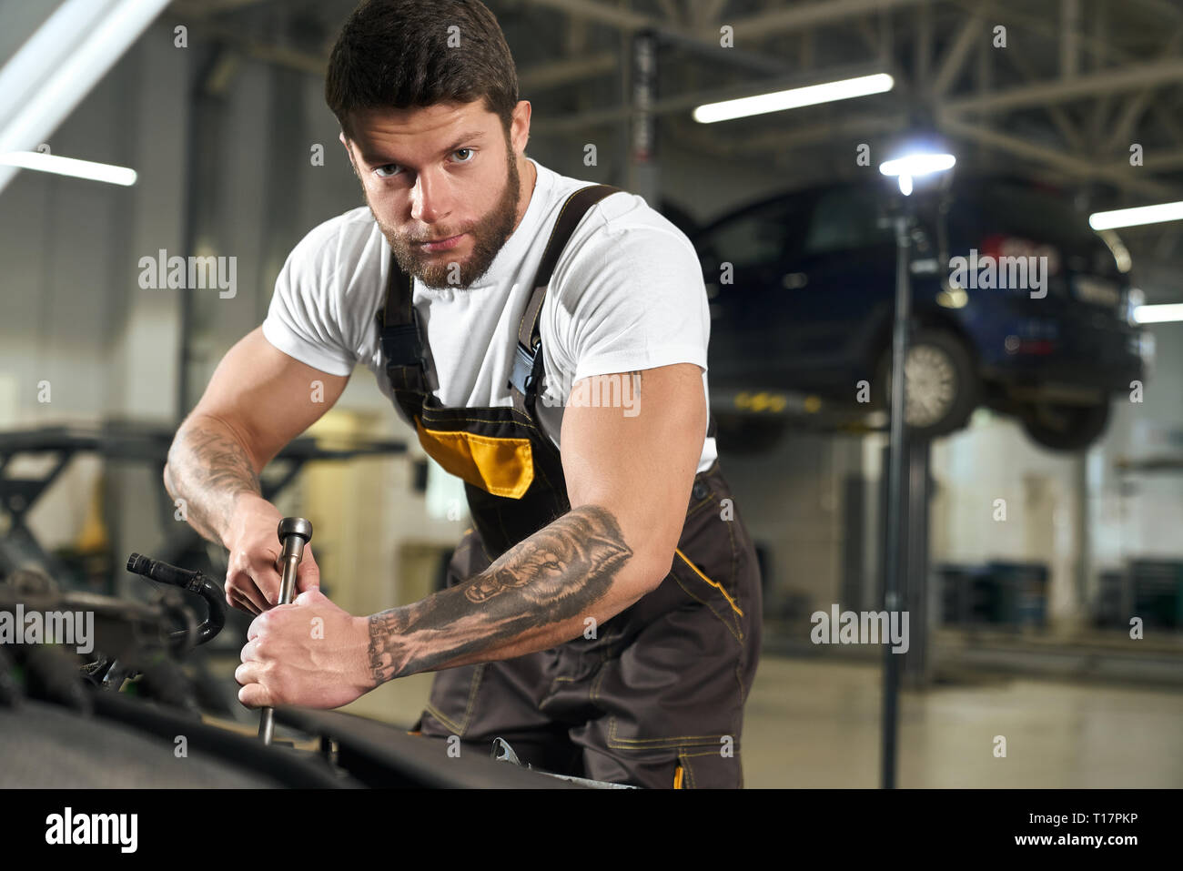 Muscular mechanic in uniform repairing car in autoservise station. Handsome, brutal bearded repairman with dark hair and tattoos on hands holding wench and looking at camera. Stock Photo