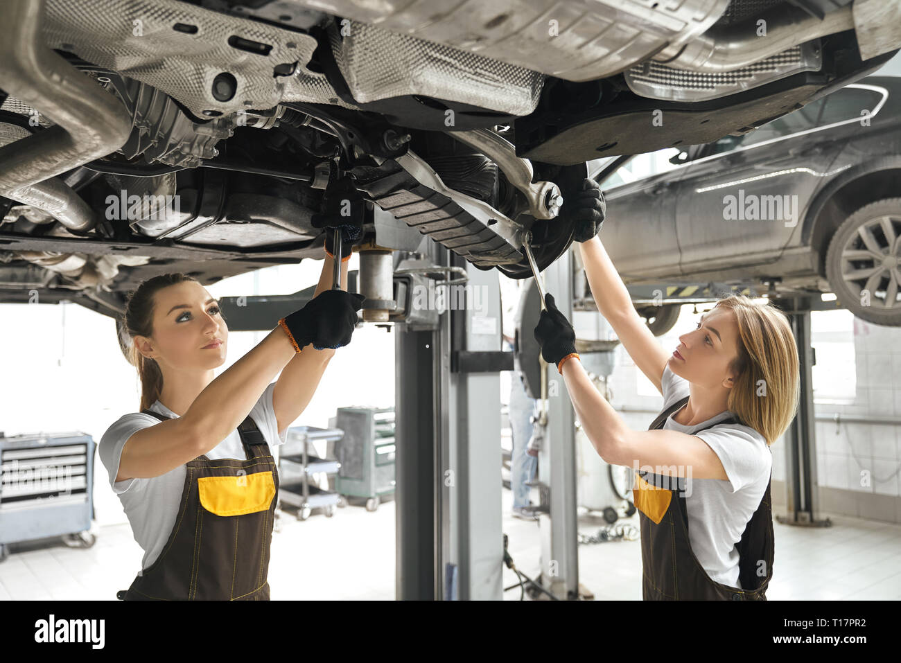 Two pretty young women in coveralls and black gloves looking up, repairing auto undercarriage. Professional female mechanics concentrated on fixing automobile. Stock Photo