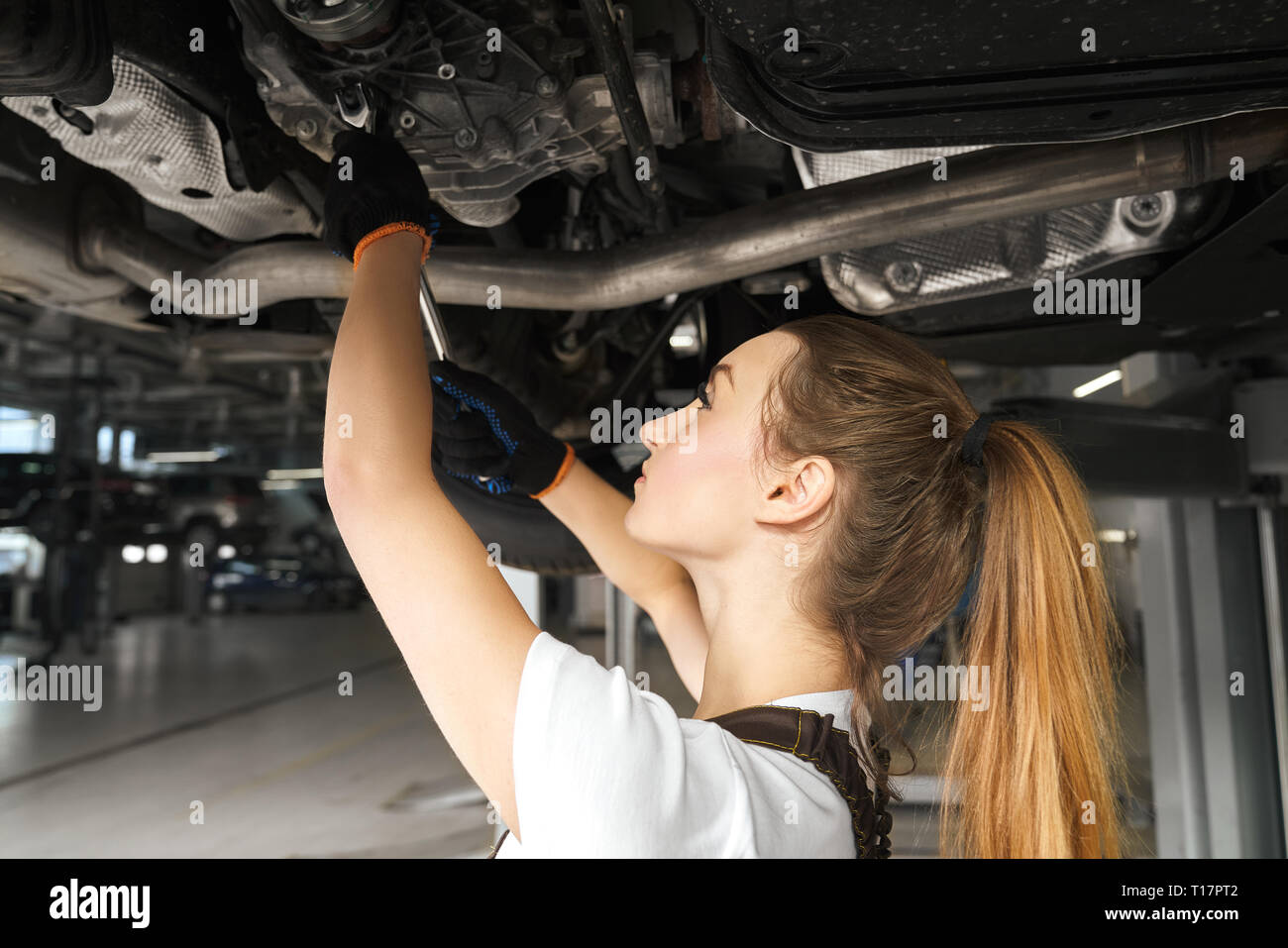 Young woman holding tool, fixing car. Girl working undercarriage of automobile, auto lifted on bridge. Female mechanic in black gloves looking up, repairing vehicle in service station. Stock Photo