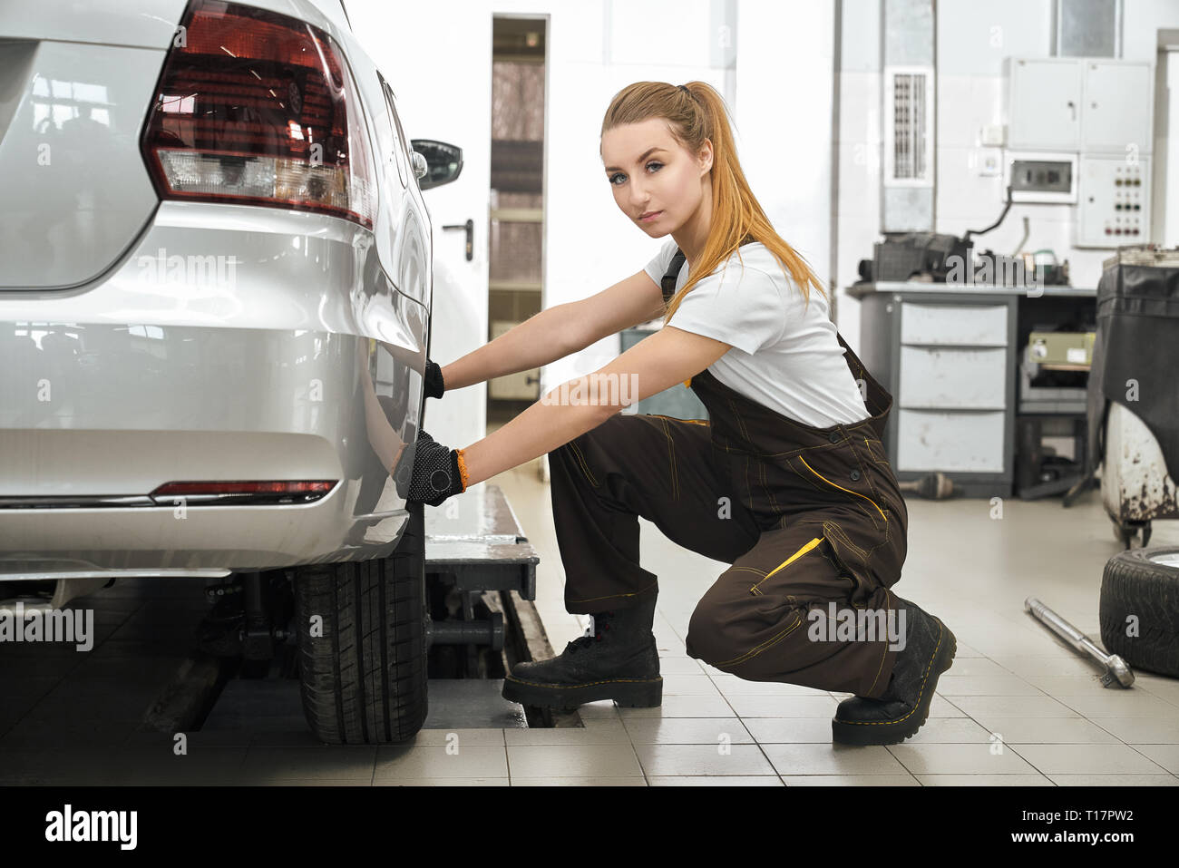 Beautiful girl fixing wheel, tyre of silver automobile. Confident young woman wearing in white t shirt and coveralls, looking at camera, posing. Girl working in service station with vehicles. Stock Photo
