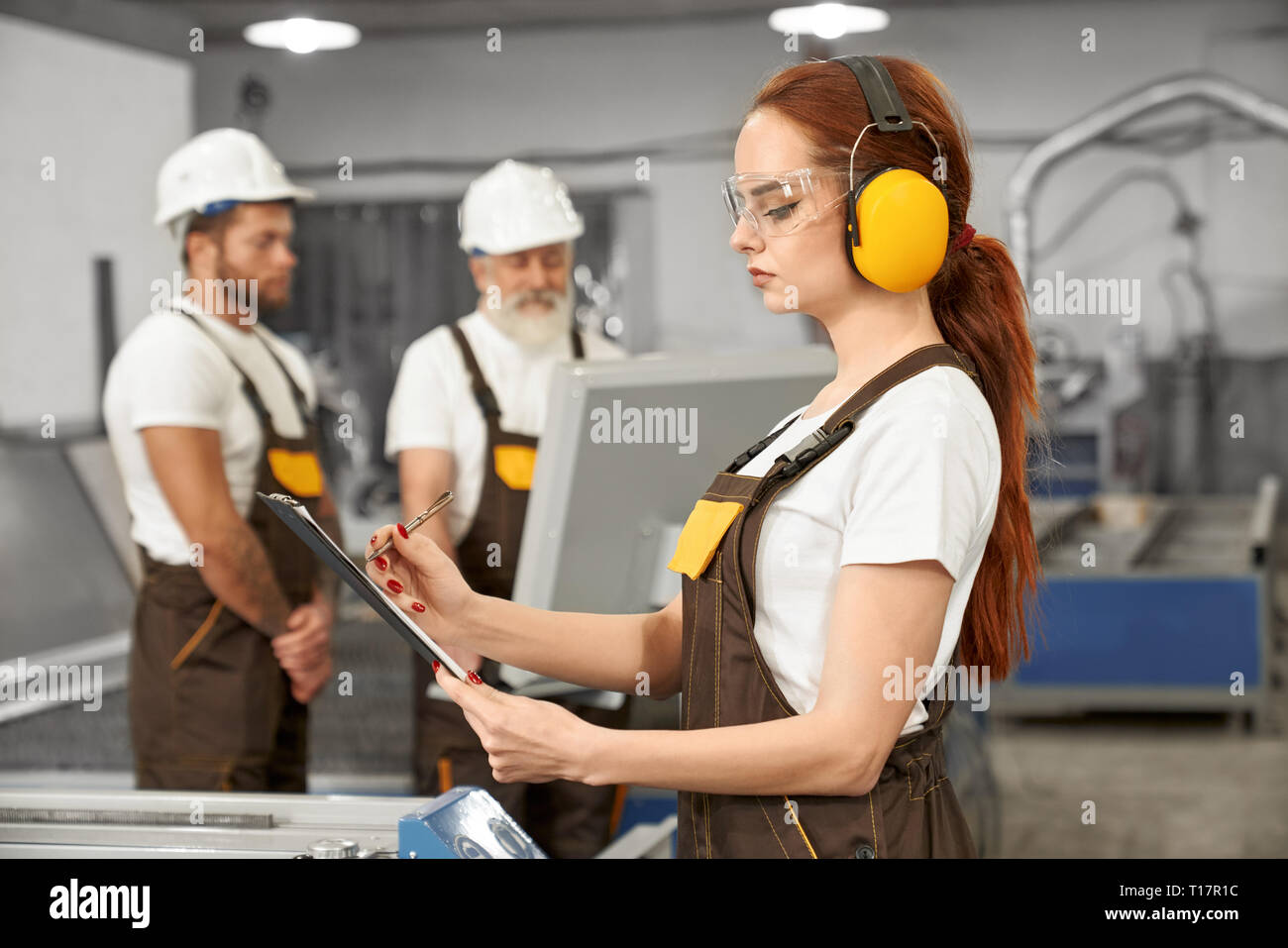 Female engineer in protective ear muffs and glasses holding folder, writing. Engineers wearing in white t shirts, coveralls, helmets. operating computerized plasma laser machine. Stock Photo