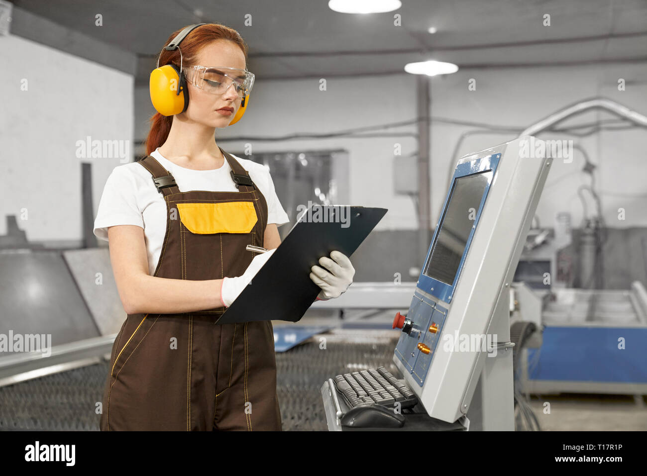 Serious, concentrated female engineer holding folder, operating computerized plasma laser machine. Beautiful woman wearing in white t shirt, gloves, coveralls, protective ear muffs and glasses. Stock Photo