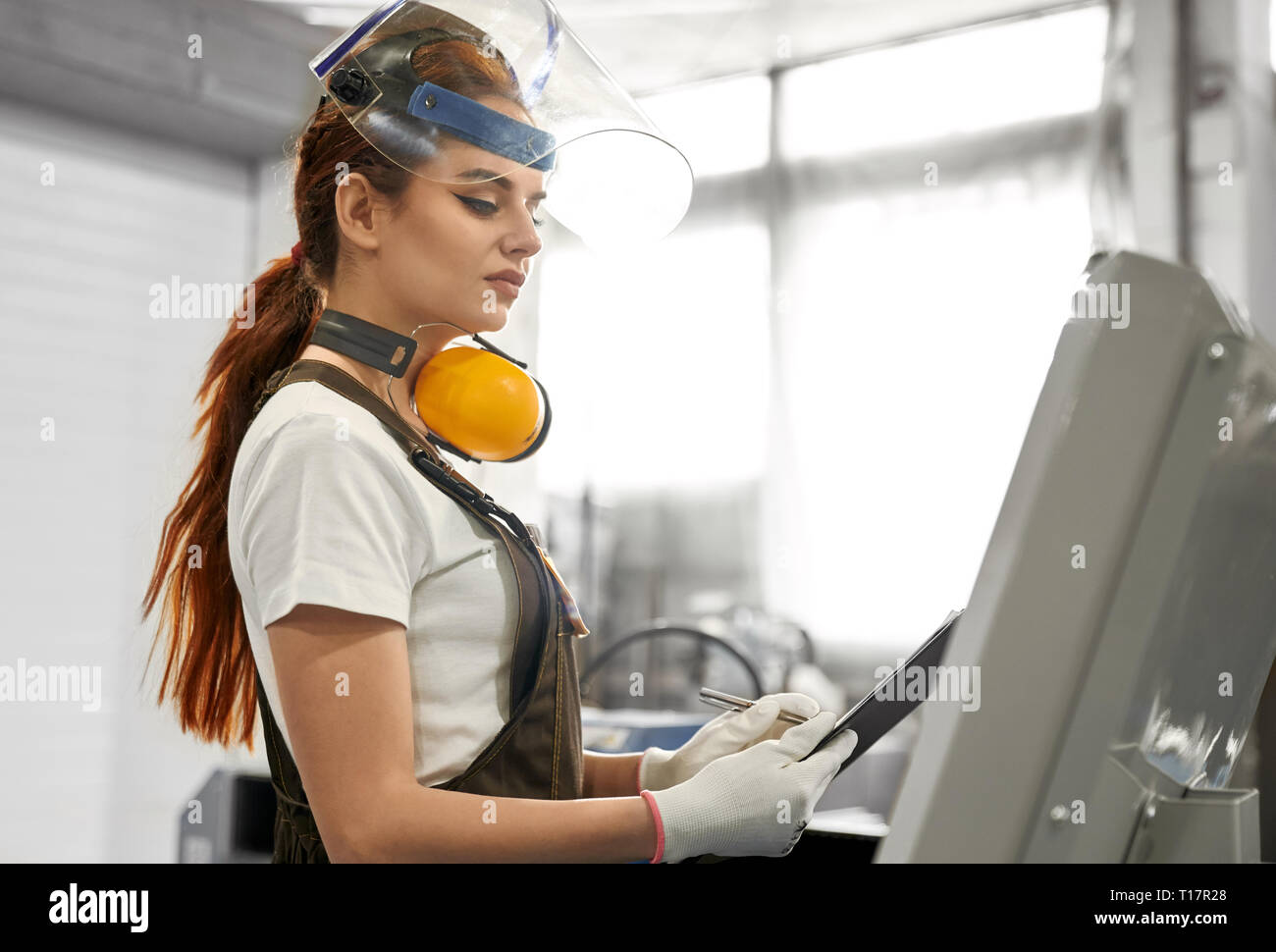 Female engineer working on factory, operating computerized machine, holding folder. Beautiful, pretty woman wearing in white t shirt, gloves, coveralls, protective ear muffs, face shield. Stock Photo