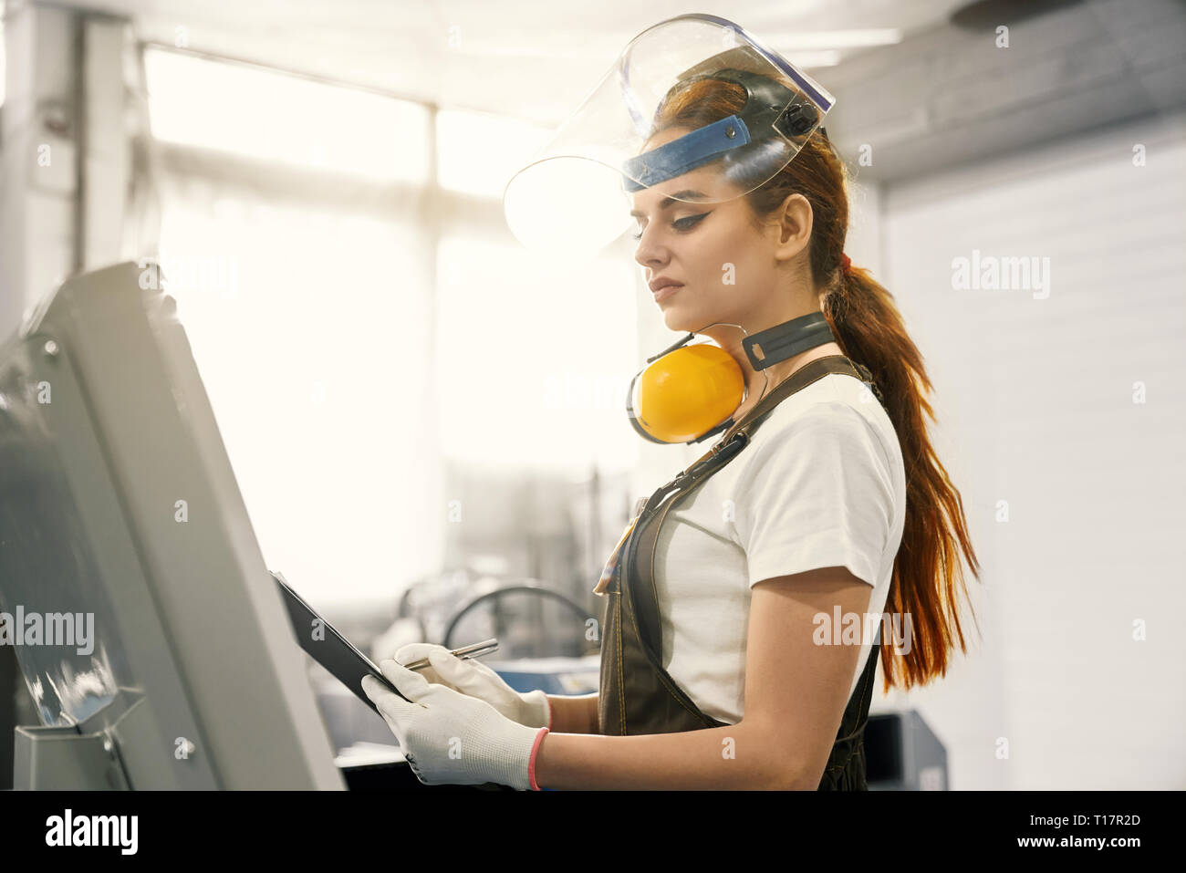Side view of beautiful woman, engineer wearing in white t shirt, gloves, coveralls, protective ear muffs, face shield. Female worker standing near computer, holding folder. Stock Photo