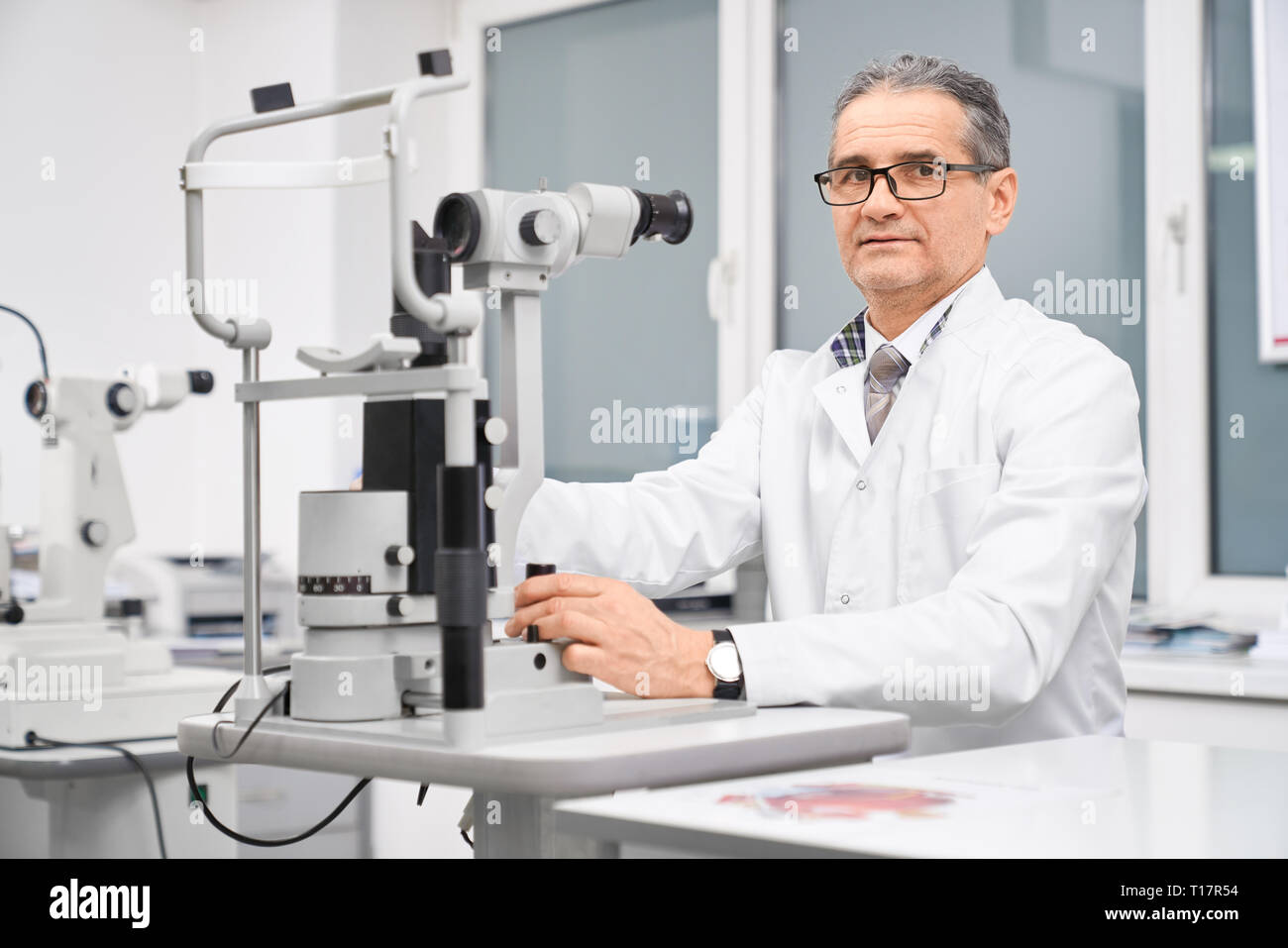 Professional doctor ophthalmologist sitting at table with special equipment slit lamp. Special device for examining eyesight of patients in medical clinic. Concept of health care and eye testing. Stock Photo