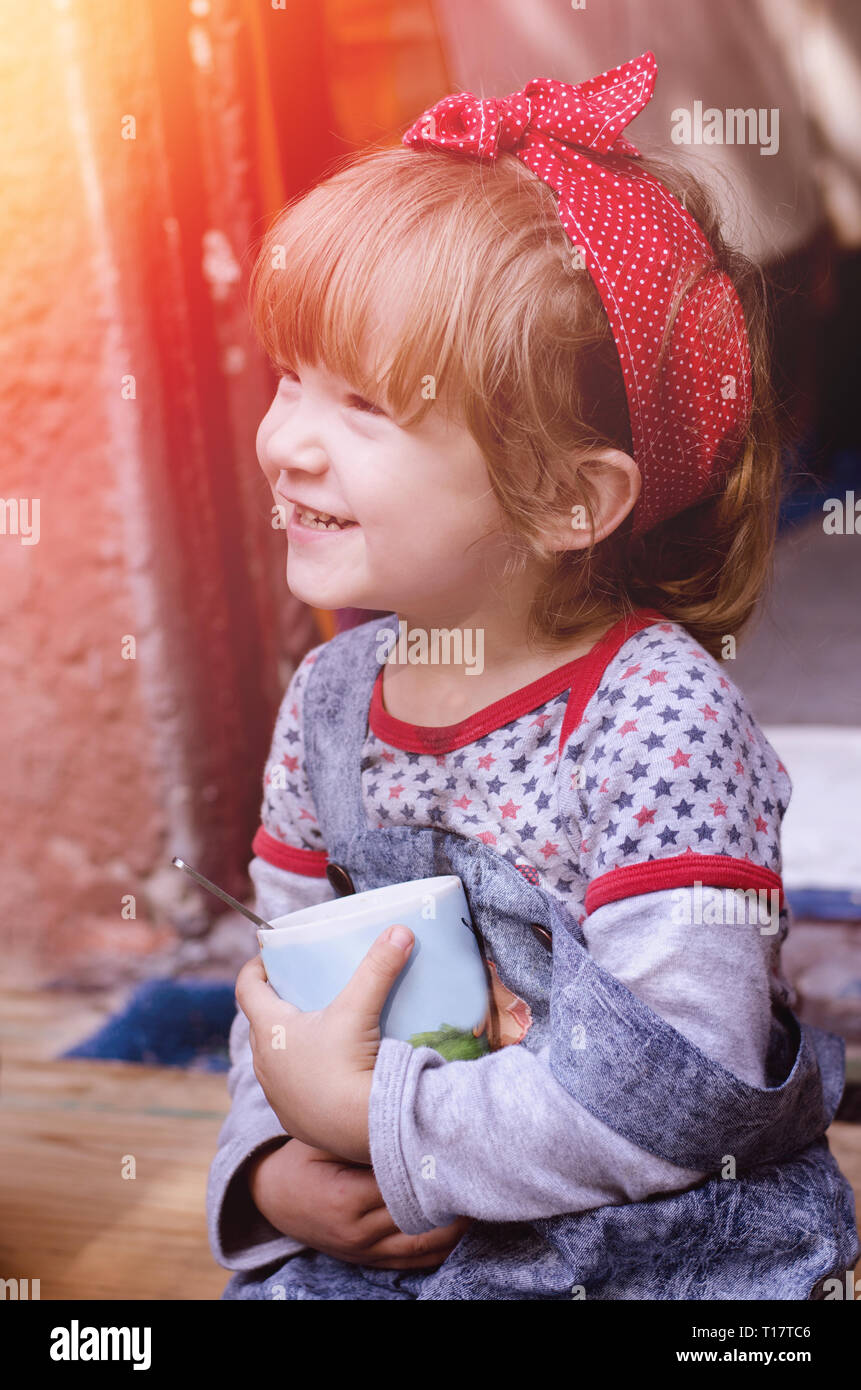 White cute toddler girl wearing bright red bandana on a had and retro style denim drinks cocoa sitting on a house threshold. Happy child portrait. Stock Photo