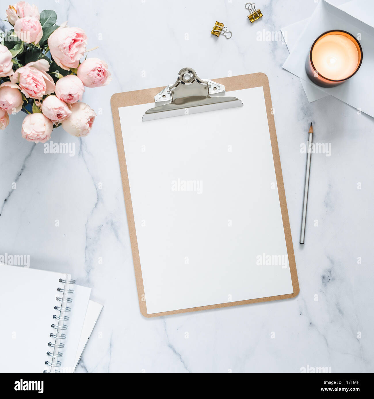 Top view of clipboard with white empty page. Clipboard, flowers, scented candle on white marble. Feminine home office mock up with blank sheet of paper A4 portrait format,copy space for text. Flat lay Stock Photo
