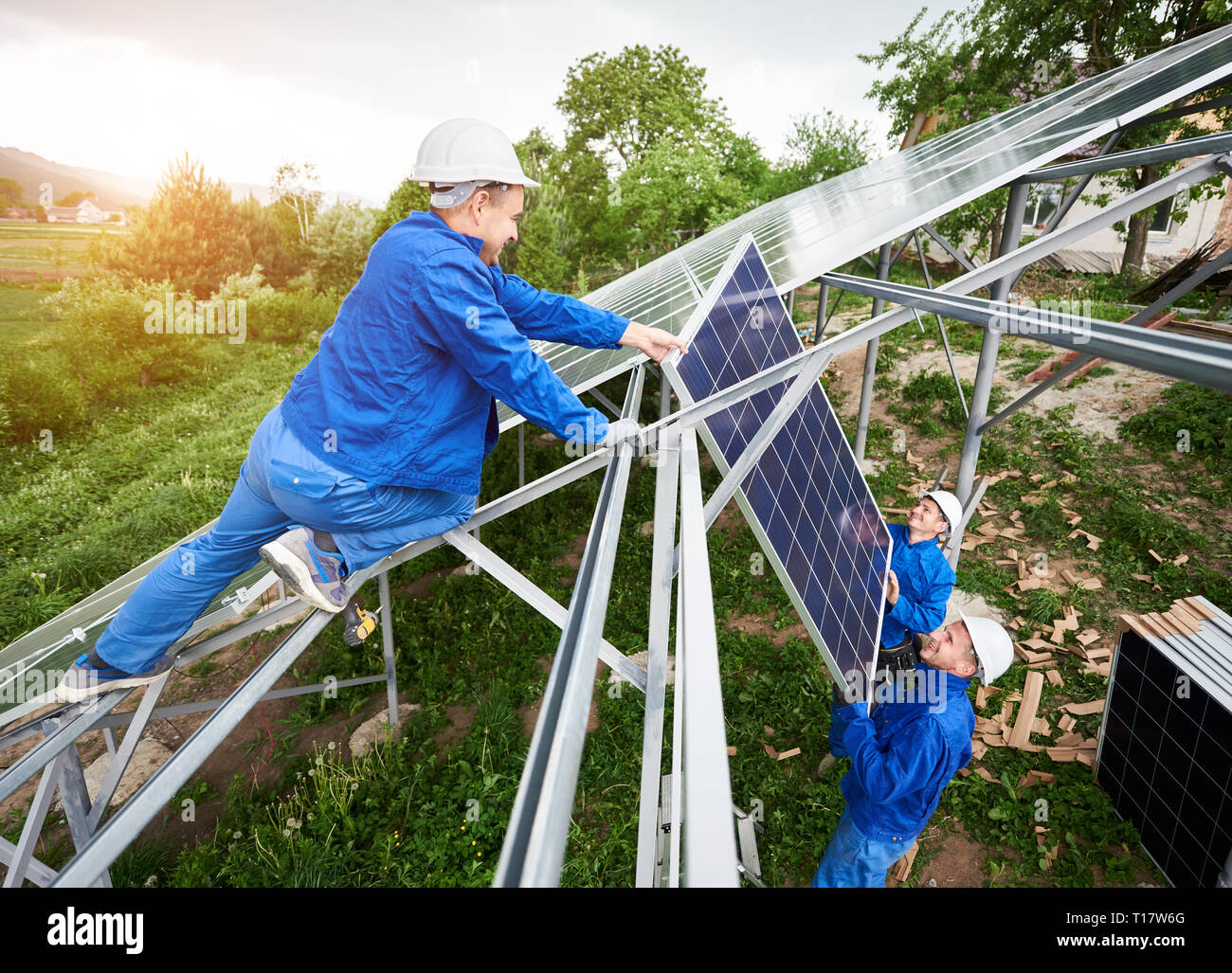 Installing of stand-alone solar photo voltaic panel system. Team of three technicians in hard-hats mounting the solar module on platform on green summ Stock Photo