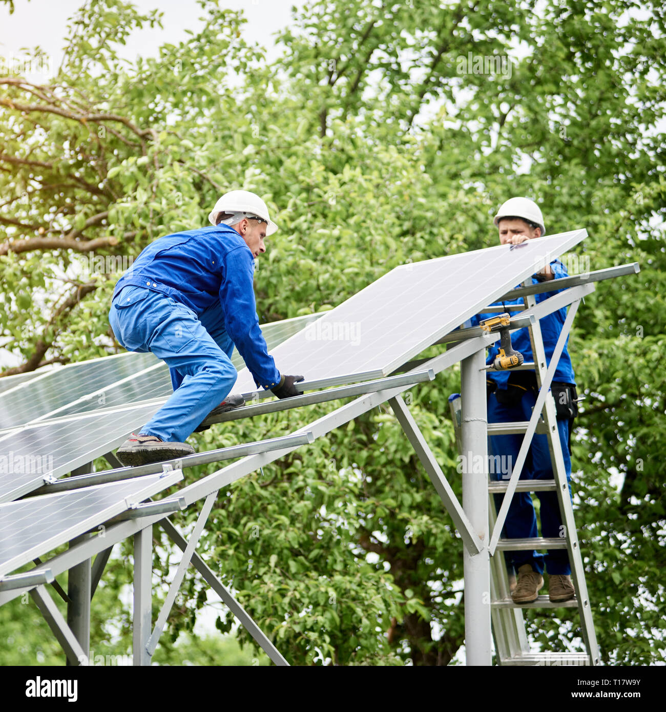 Installing of stand-alone solar photo voltaic panel system. Two workers assembling solar modules on metal platform on green tree background. Alternati Stock Photo