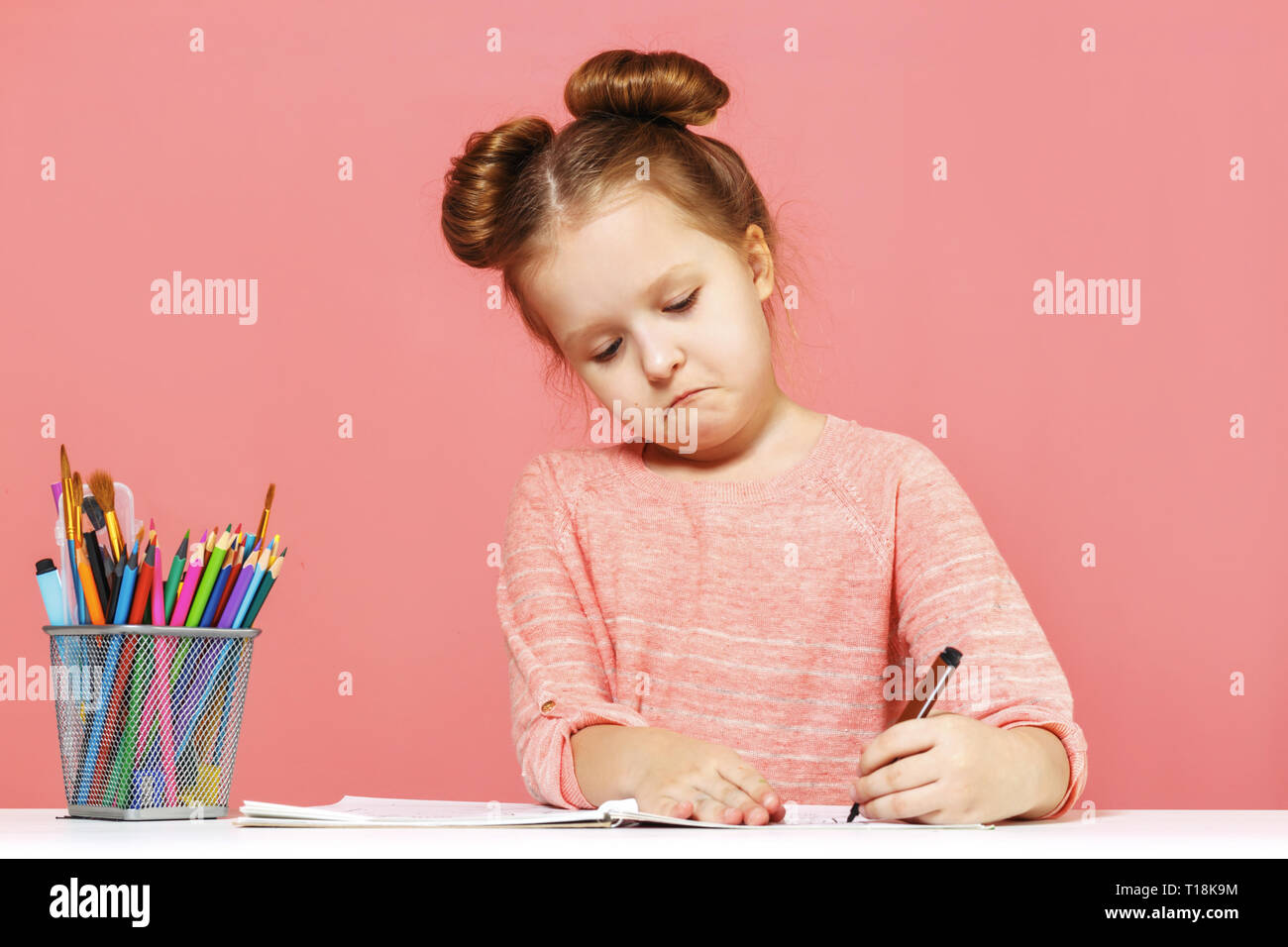 A cute little baby girl is sitting at the table and drawing with enthusiasm. Pink Background Close-Up Stock Photo