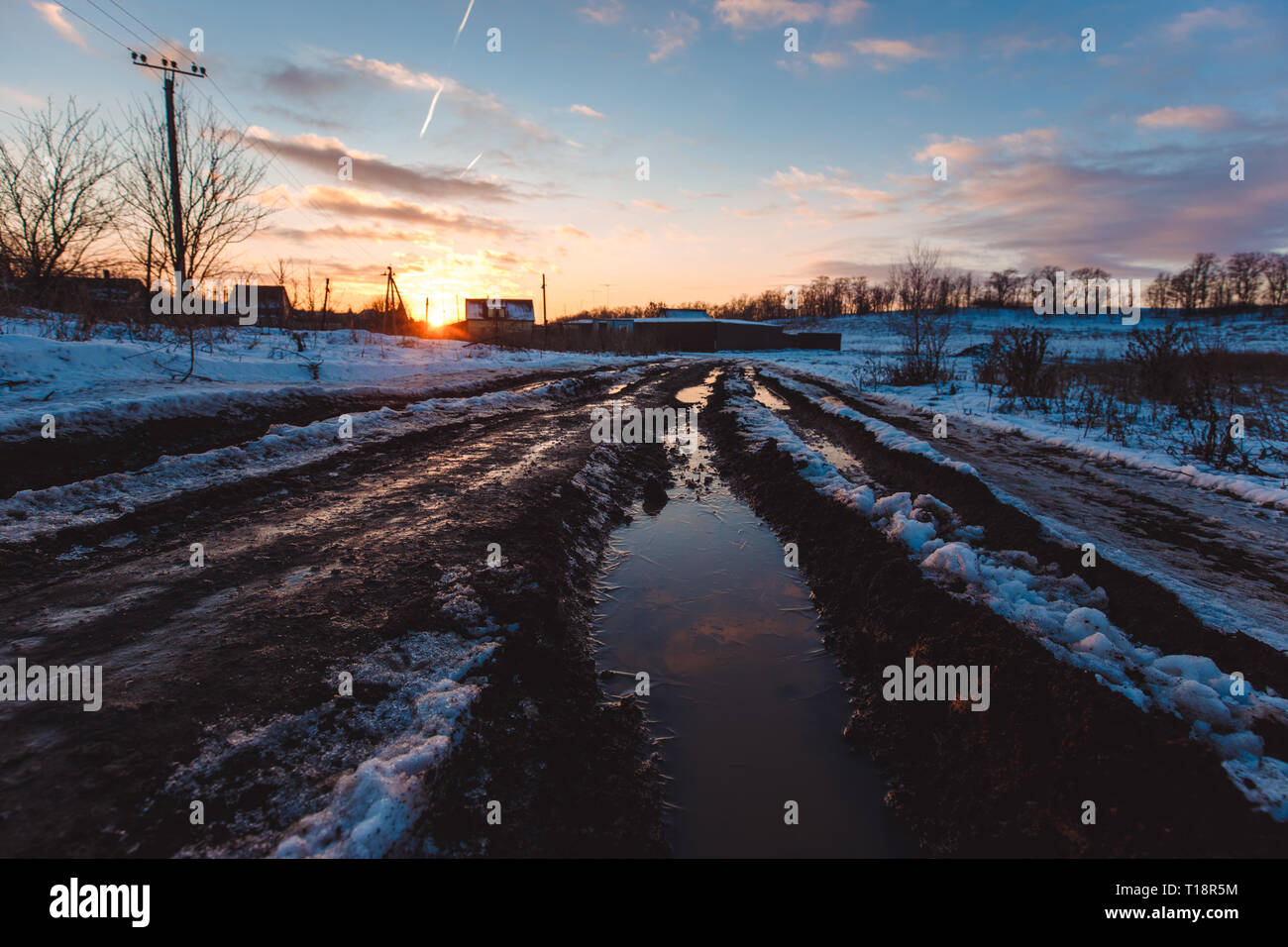 Country dirt road with muddy puddles and melting snow in early spring at sunset Stock Photo